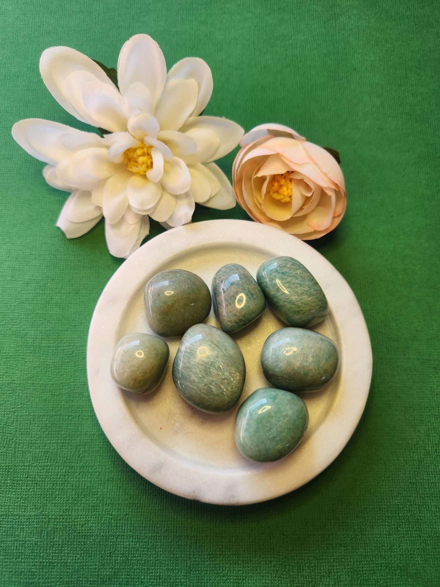 Amazonite tumble stones displayed in a white dish, with soft flowers in the background and a green backdrop for a calming, natural look.