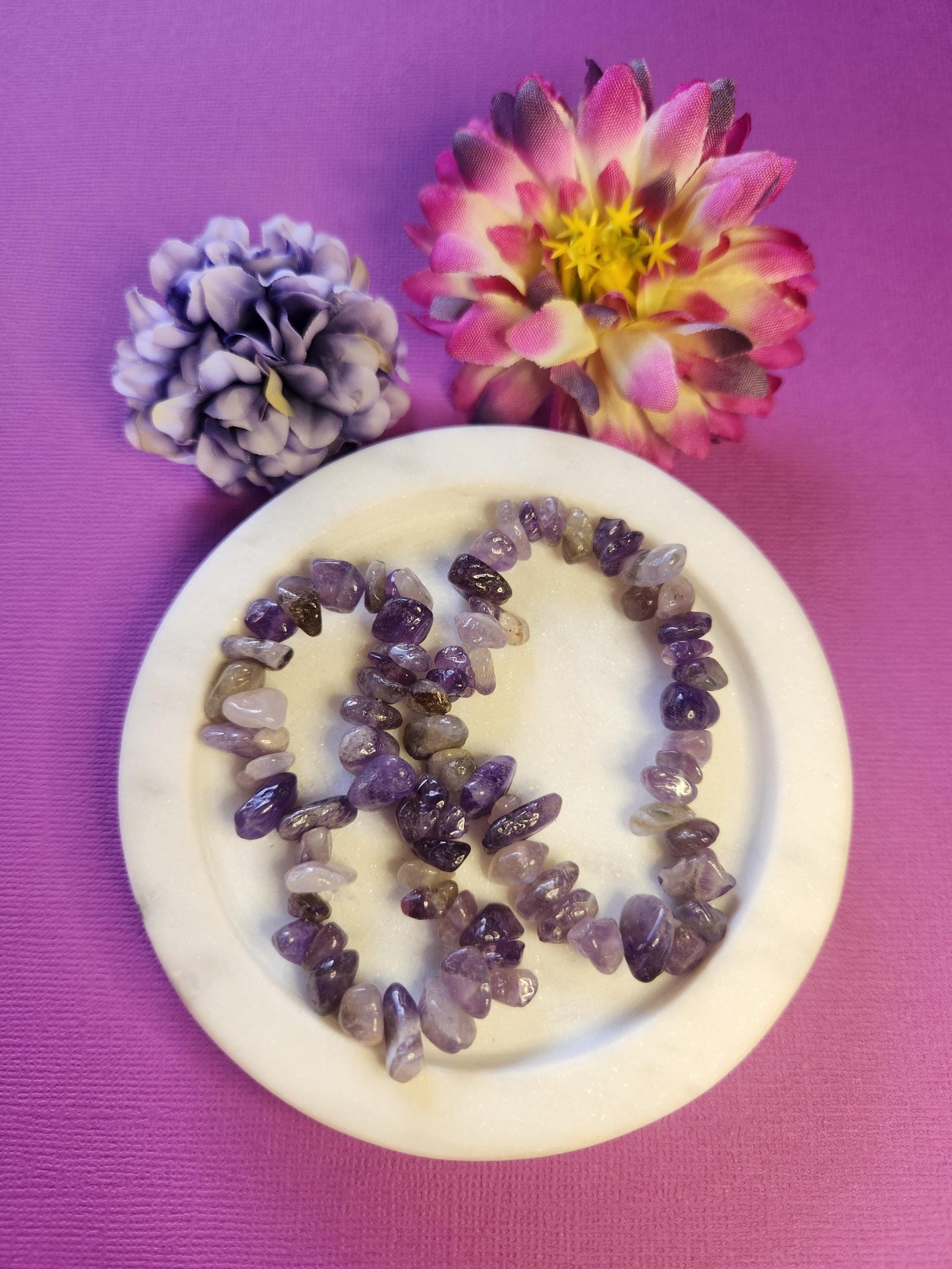 Two amethyst chip bracelets displayed in a white dish with two flower props on a purple backdrop, showcasing natural purple crystal chips that promote calm, intuition, and spiritual healing—perfect crystal jewelry gift for meditation and energy balance.