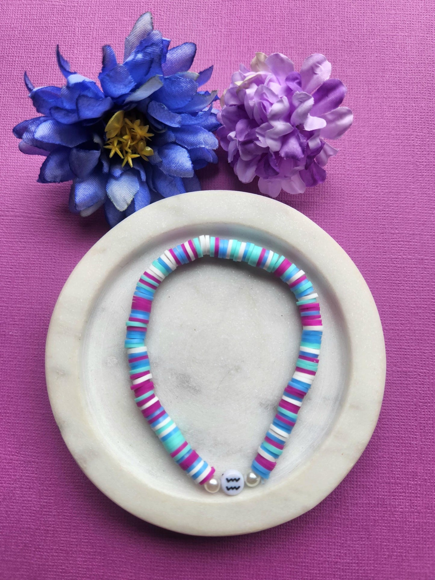 Aquarius zodiac clay beaded bracelet in white dish with two flower props on purple background, handmade light blue purple white mint beaded astrology bracelet with Aquarius symbol bead, stretchy elastic zodiac jewellery product photo