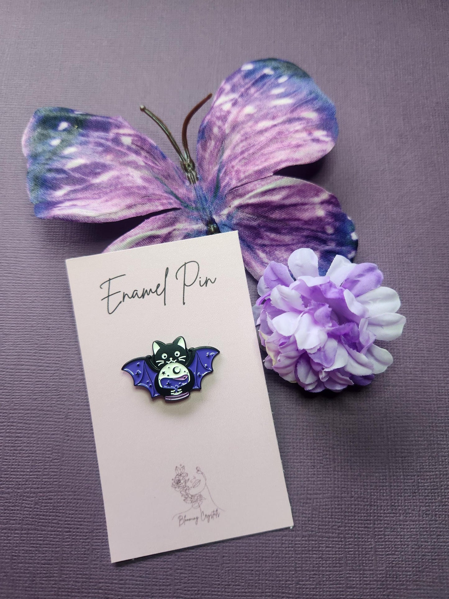 Cute black cat enamel pin with purple bat wings, holding a silver-outlined crystal ball. Displayed with a purple butterfly and flowers on a purple background. Spooky cute pin, gothic cat enamel pin, bat cat witchy accessory, Halloween lapel pin, mystical familiar aesthetic.