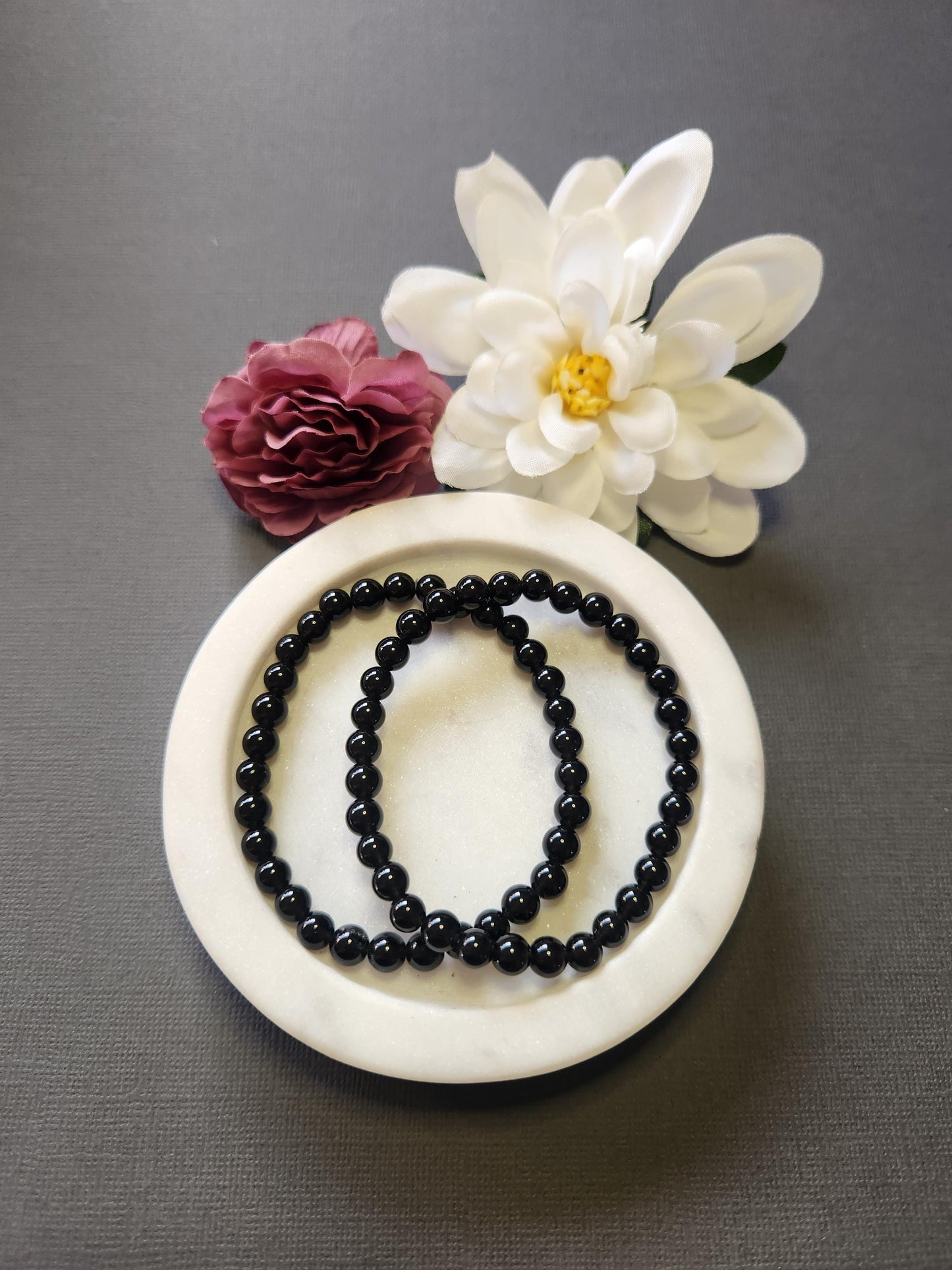 Photo showcasing two Black Obsidian 6mm beaded bracelets in a white dish with two flower props on a dark grey background, highlighting the polished black crystal beads, grounding energy, and protective properties of Obsidian — perfect for spiritual healing and energy shielding.