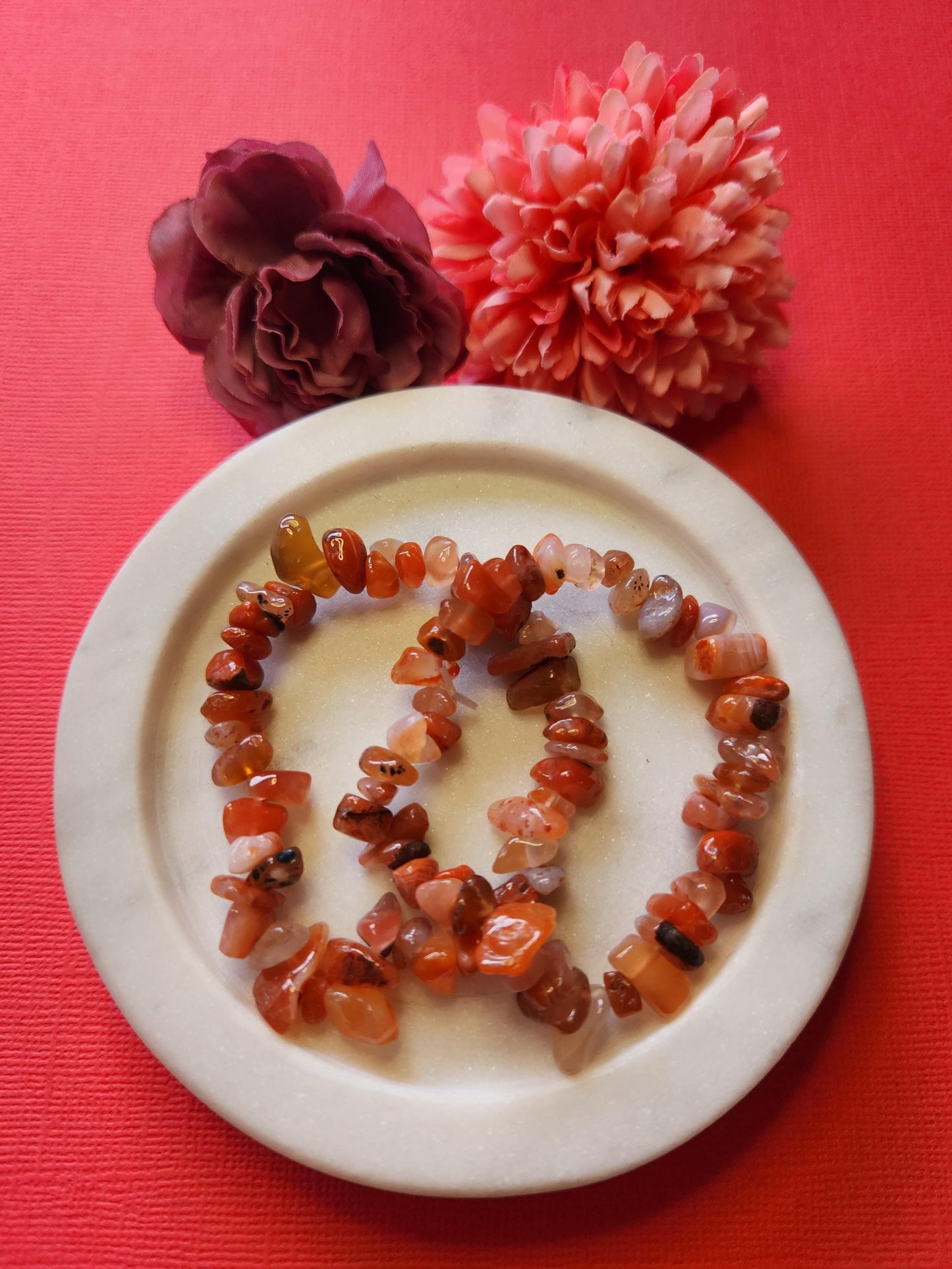 Two Carnelian chip stretch bracelets displayed on a white dish with two flower props, set against a bright red background. Natural carnelian gemstones for energy, motivation, and emotional balance. Perfect for crystal healing, chakra work, meditation, or as a thoughtful gift.