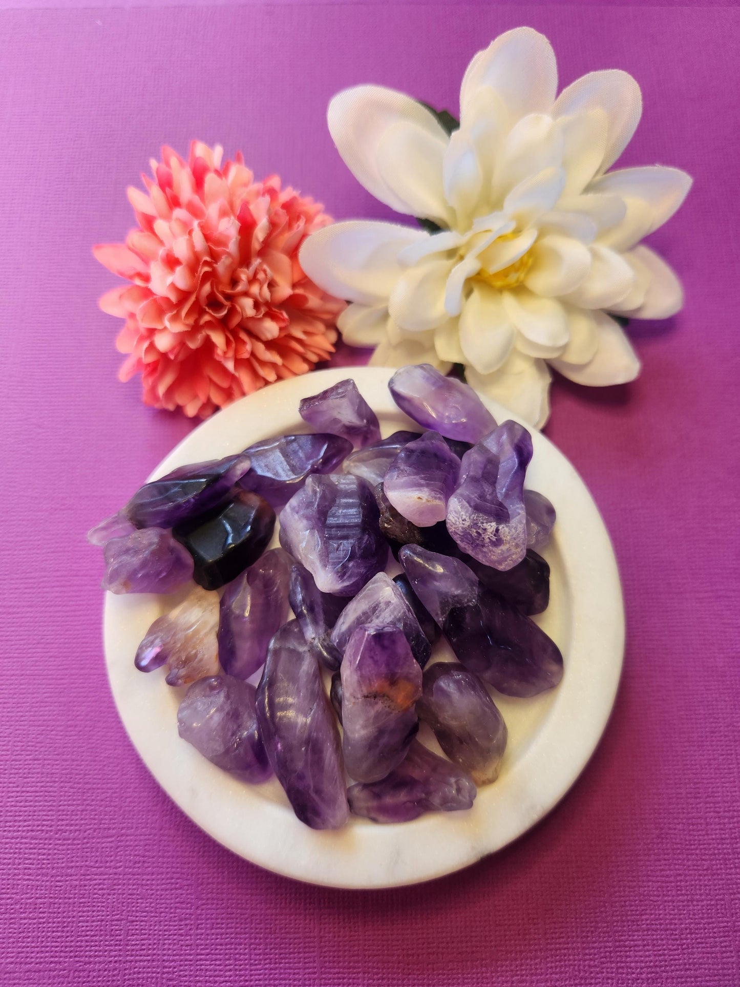Chevron Amethyst rough tumble displayed on a white dish with flower props in the background, set against a purple backdrop, highlighting its unique banded purple and white crystal patterns.