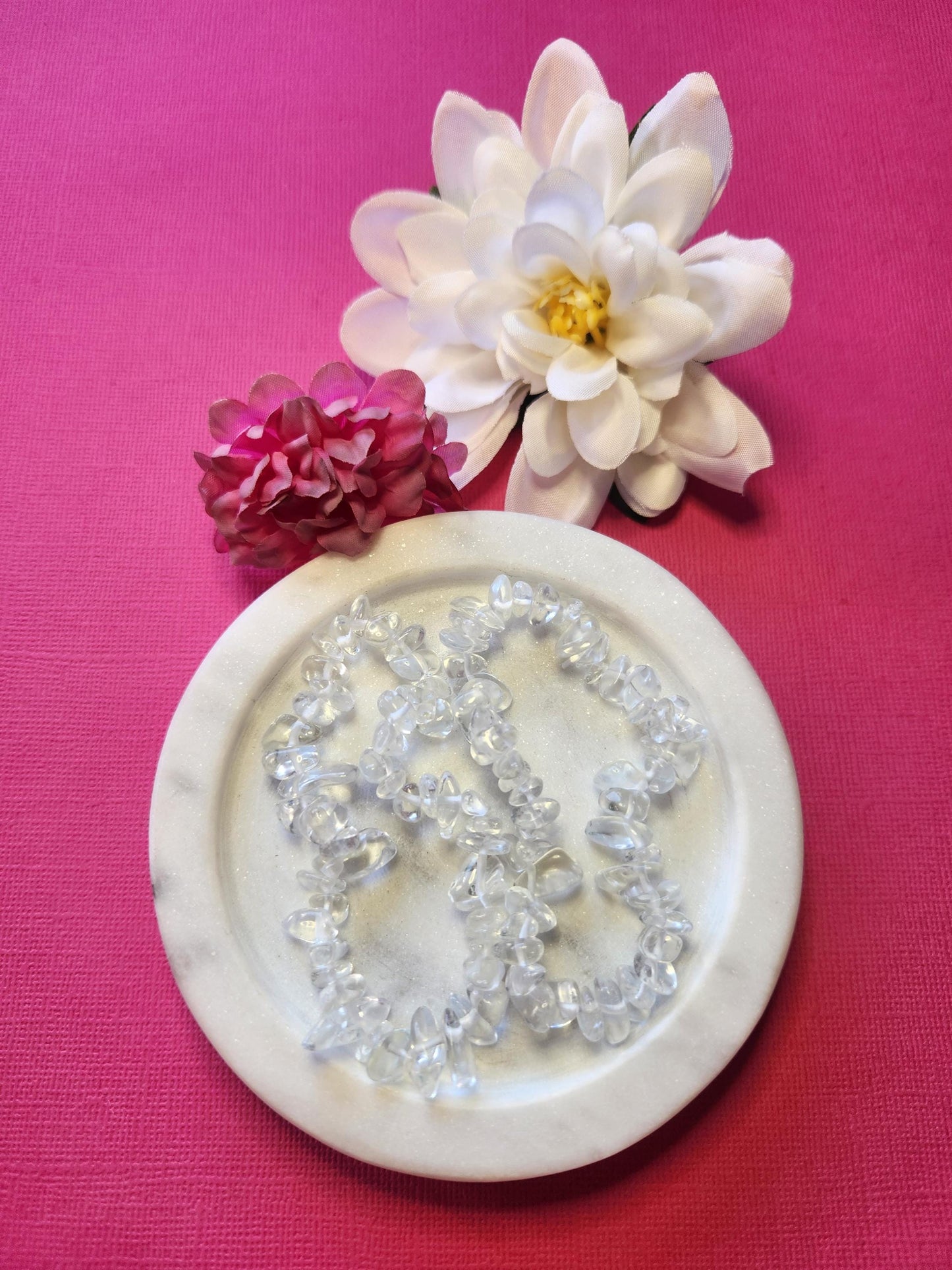 Two Clear Quartz chip bracelets displayed in a white dish with two flower props on a bright pink background, featuring polished clear crystal chips that shimmer with light energy — perfect clarity and healing crystal jewelry for focus, balance, and spiritual cleansing.