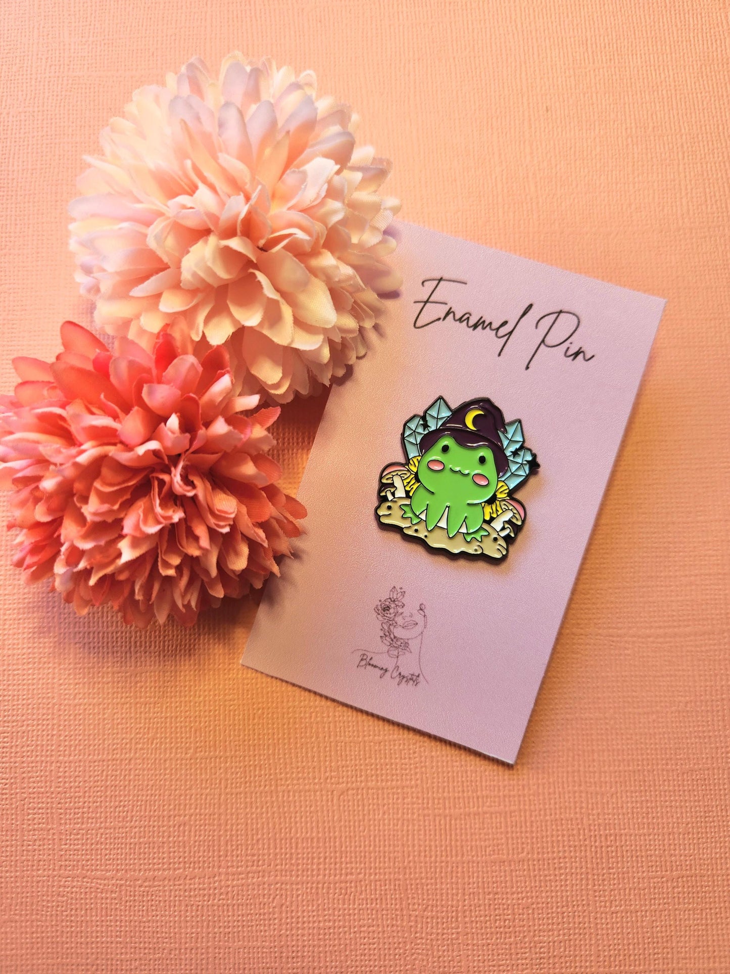Enamel pin featuring a cute green frog wearing a purple witch hat, surrounded by crystal designs, displayed on a light pink background. This whimsical frog witch pin blends magical and cottagecore aesthetics, making it a charming accessory for bags, jackets, or pin boards. The soft pink backdrop enhances the vibrant colors of the enamel design, perfect for collectors of witchy, fantasy, or frog-themed accessories.