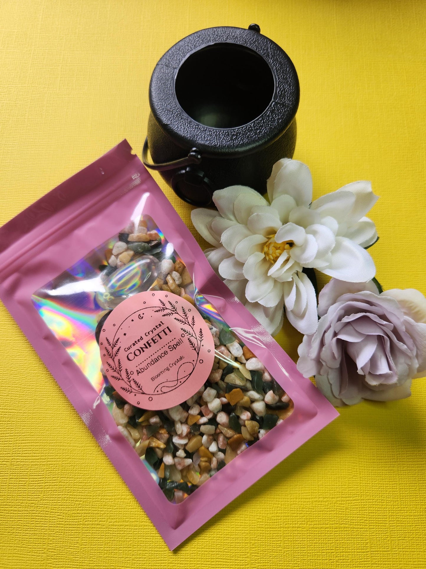 Abundance spell crystal confetti packaged in a shiny pink holographic bag with a label. Includes crystal chips—Yellow Aventurine, Sunstone, Green Aventurine—and tumbled stones of Moss Agate, Clear Quartz, and Amazonite. Comes with a black cauldron and is styled with two flower props on a bright yellow background. Prosperity crystal mix, money spell kit, abundance ritual stones, witchcraft confetti for manifestation.