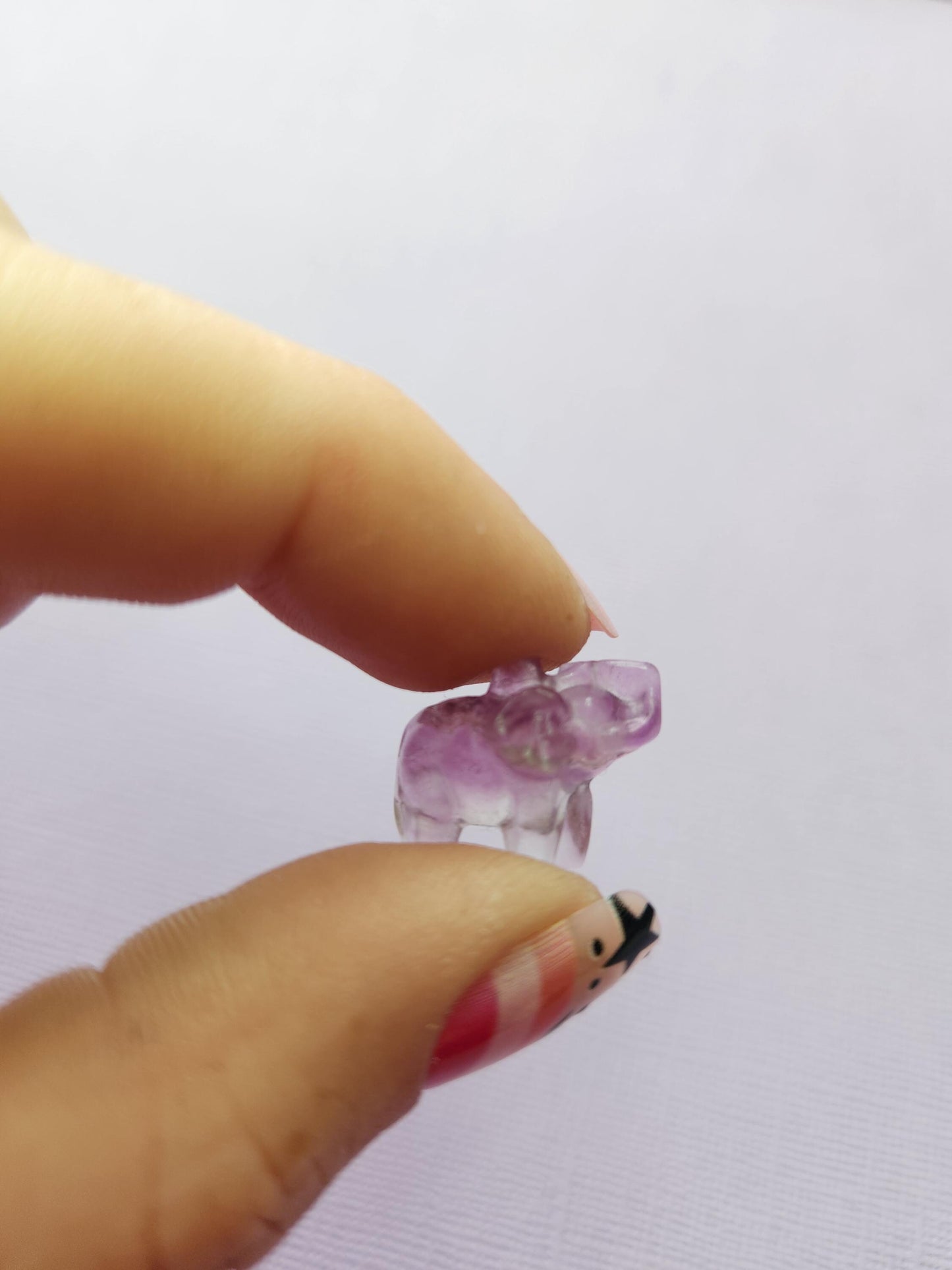 Close-up photo of a single fluorite mini elephant carving held between fingers, showing polished details and natural purple, green and blue fluorite banding against a lavender backdrop. Ideal for fluorite carvings, mini crystal animals, elephant crystal collectors and pocket-sized gemstone companions