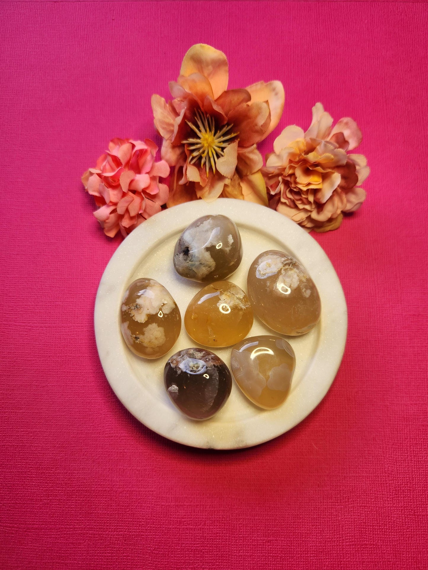 Flower Agate tumbles displayed on a bright pink background, showcasing their soft, translucent tones with natural floral-like inclusions. These polished gemstones are known for supporting emotional healing, self-growth, and feminine energy. Ideal for meditation, self-love rituals, and spiritual work. The vibrant pink backdrop enhances the gentle beauty of the stones, making them perfect for gifting or adding to a crystal collection.