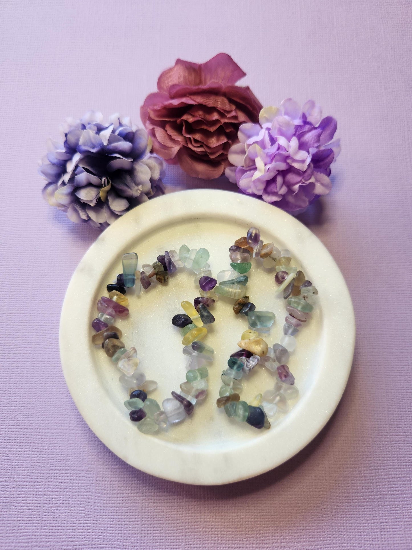 Two Fluorite chip bracelets displayed in a white dish with three flower props on a lavender background, featuring polished purple, green, and clear crystal chips that promote focus, mental clarity, stress relief, and chakra balancing — perfect healing crystal jewelry gift.