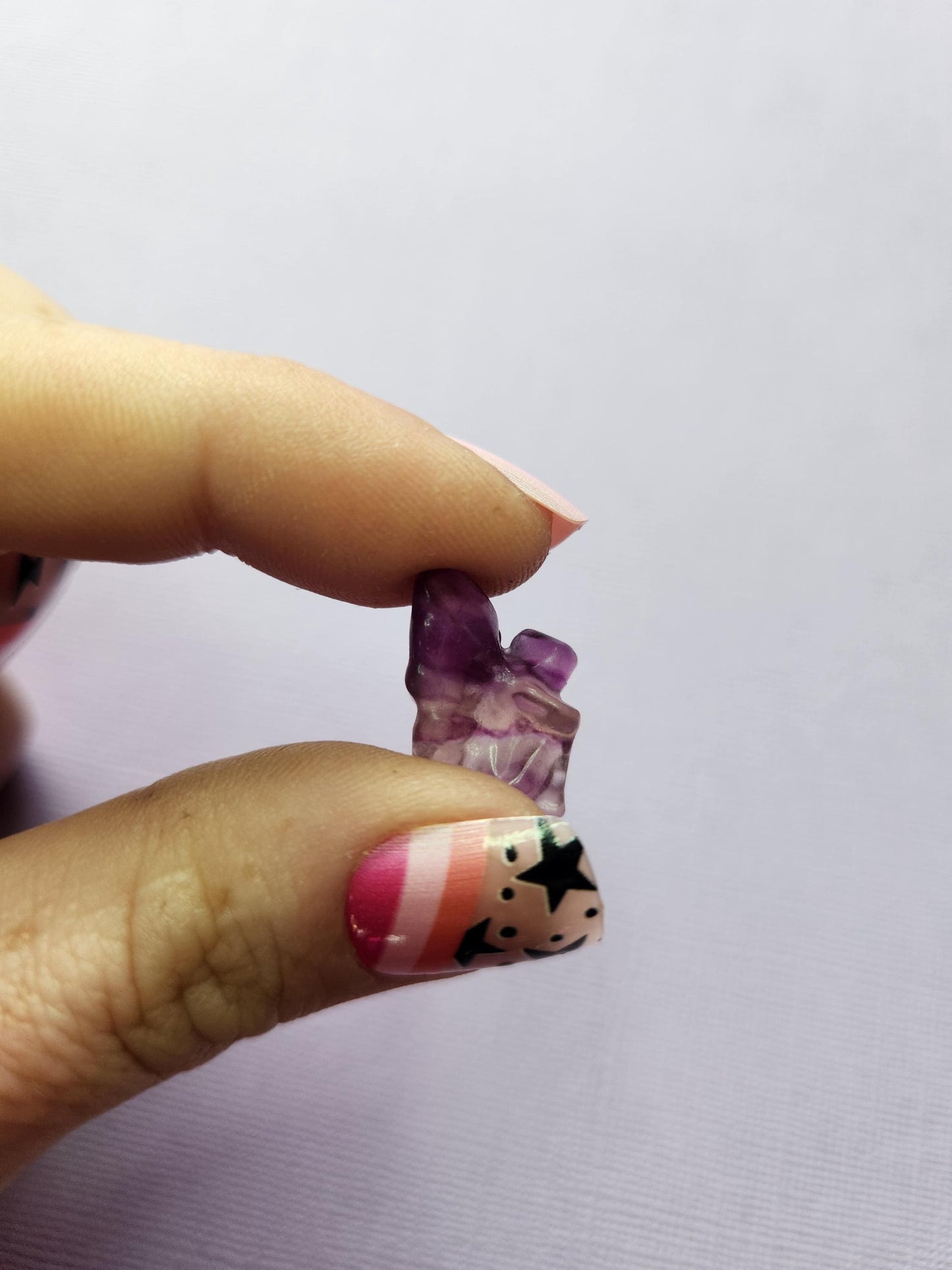 Close-up photo of a single fluorite mini fairy carving held between fingers, showing polished details and natural purple, green and blue fluorite banding against a lavender backdrop. Perfect for fluorite carvings, mini crystal fairies, collectible gemstone figures and pocket-sized magical crystals