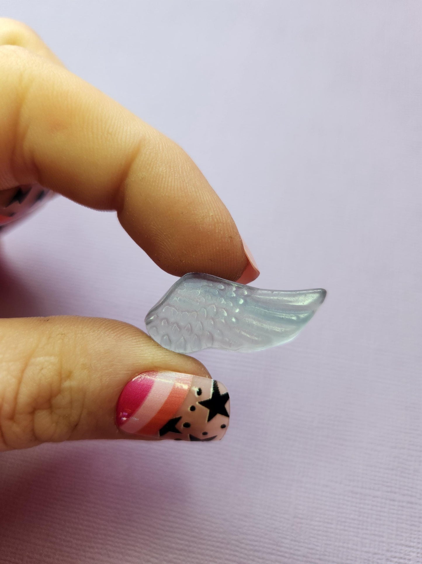 Close-up photo of a single fluorite mini wing carving held between fingers, showing smooth polished details and natural purple, green and blue fluorite banding against a lavender backdrop. Perfect for fluorite carvings, angel wing crystals, collectible gemstone accents and pocket-sized crystal pieces.