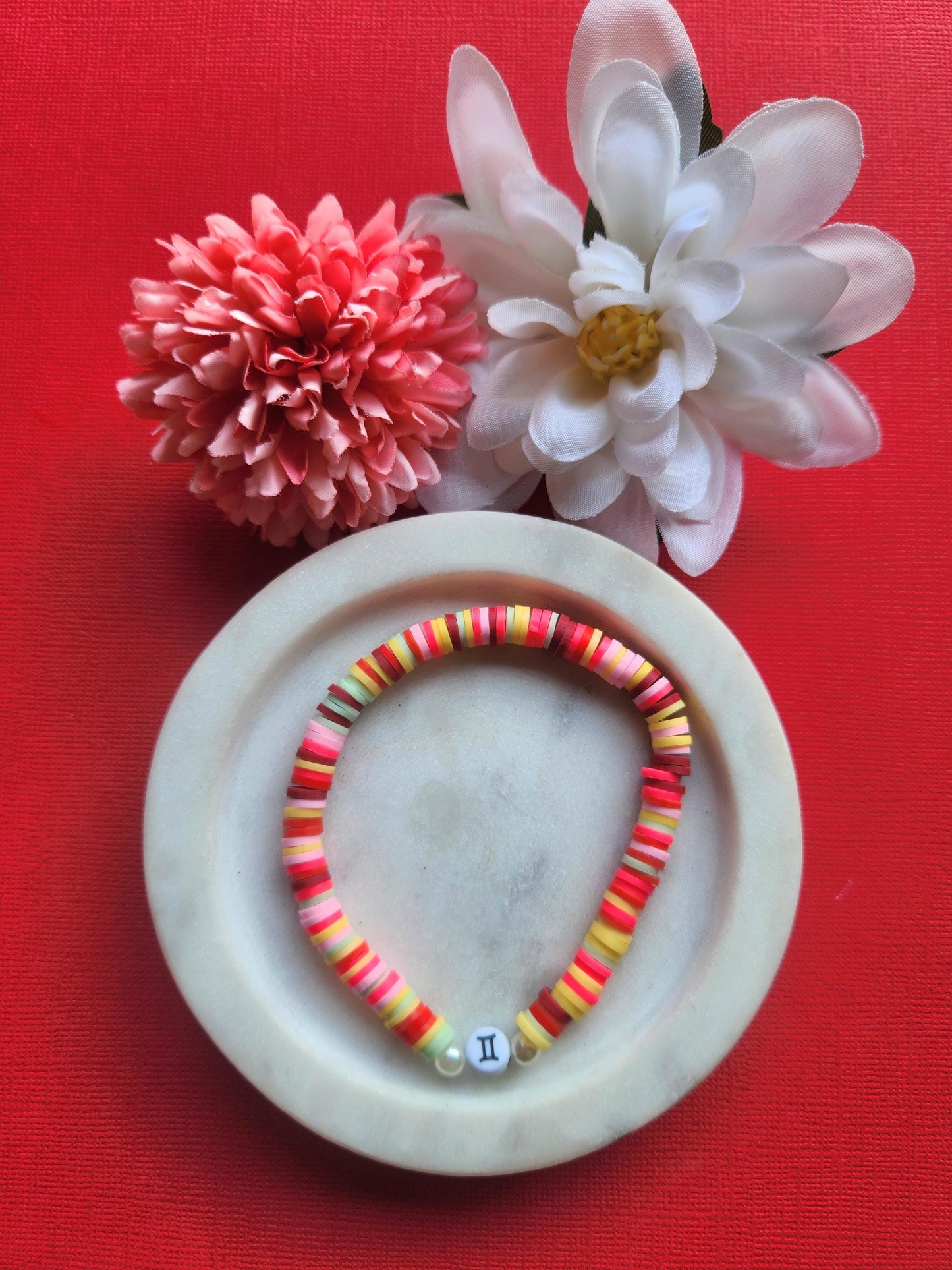 Gemini zodiac clay beaded bracelet in white dish with two flower props on red background, handmade pink red maroon yellow and white polymer clay beads with Gemini symbol bead, elastic stretch astrology jewellery product photo