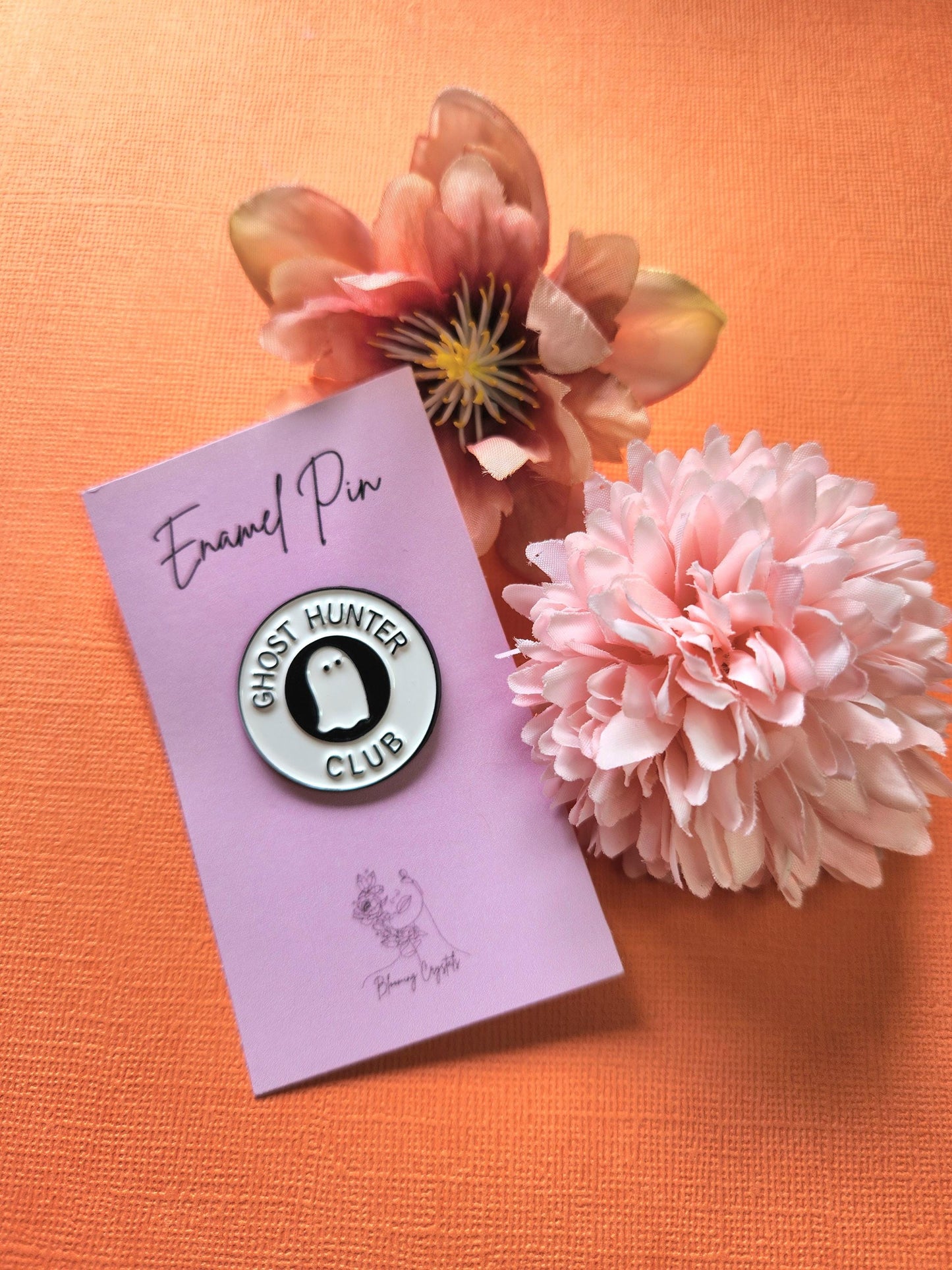 Round white enamel pin with a black center featuring a white ghost illustration. The outer border reads "Ghost Hunter Club" in black text. Styled with two flower props on a bright orange background. Cute ghost pin, paranormal enamel pin, ghost hunting club aesthetic, spooky lapel pin, Halloween accessory for ghost lovers.