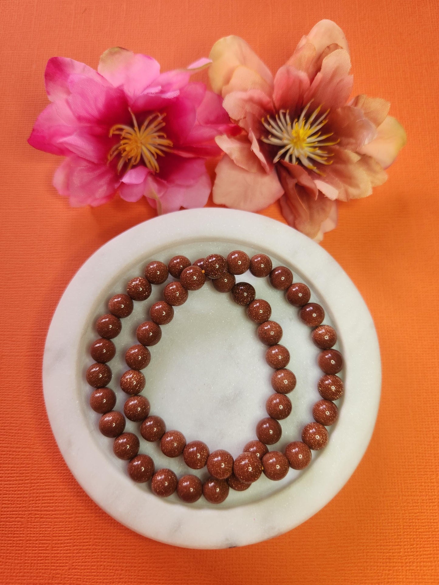 Two 8mm Gold Sandstone beaded bracelets in white dish with two flower props on orange background, natural gold sandstone gemstone beads, crystal jewellery, handmade beaded bracelet for grounding, protection and energy healing, gemstone accessory product photo