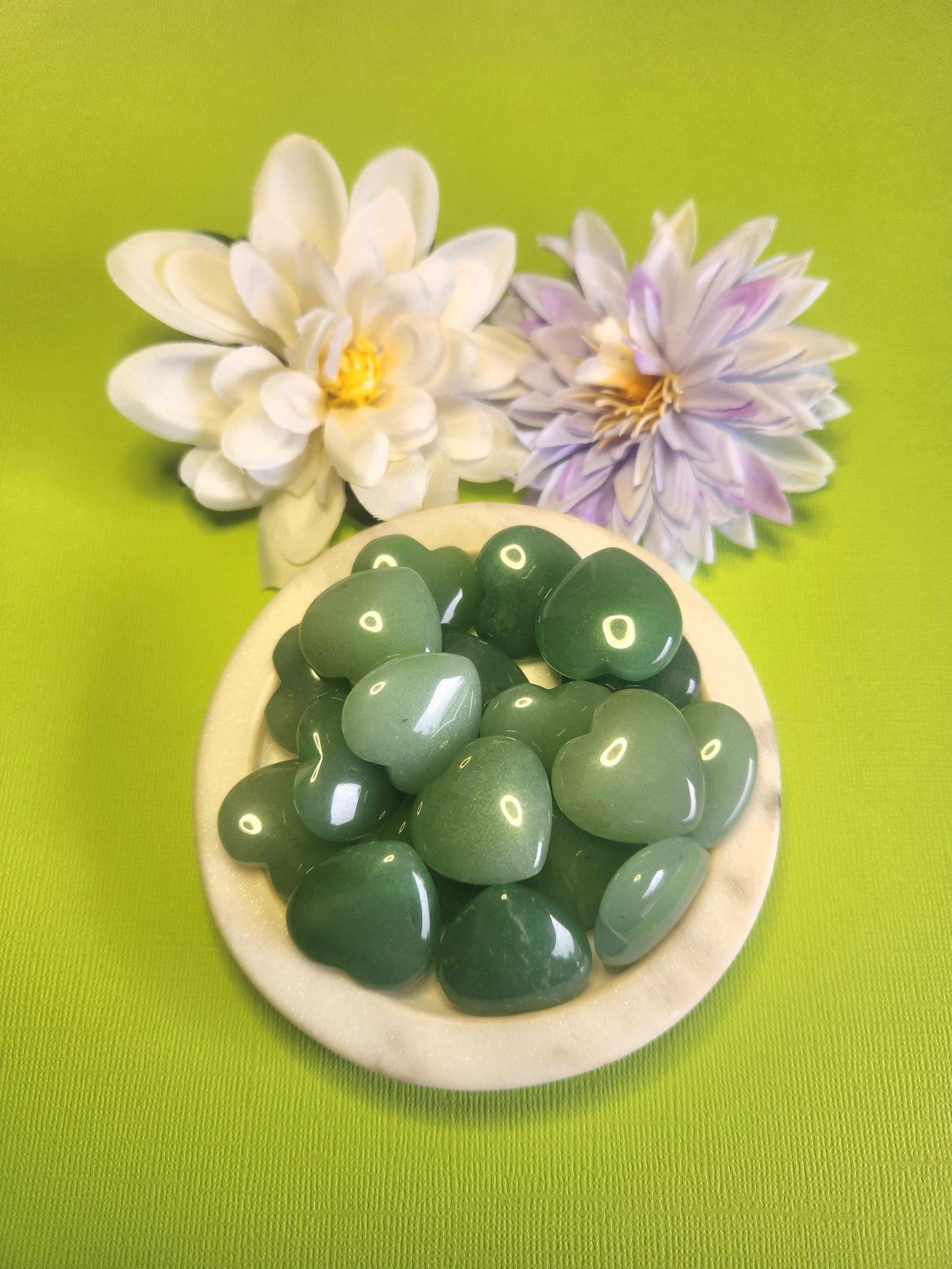Green aventurine crystal heart displayed in a white dish on a green background with soft flower props featuring polished heart stone for luck abundance love healing crystal decor and gift