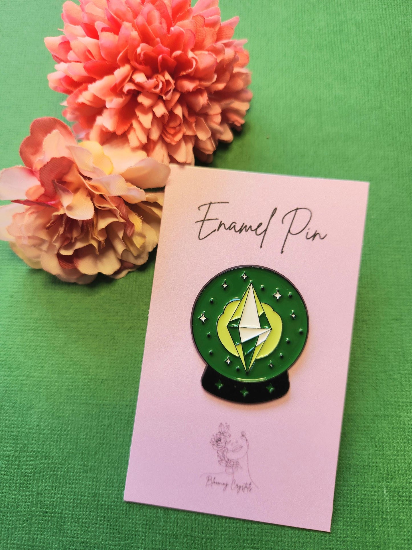 Green and black crystal ball enamel pin with a green and yellow diamond-shaped crystal inside, displayed on a pink backing card with two flower props on a green background, mystical witchy aesthetic enamel pin for bags jackets gifts and pin collectors.