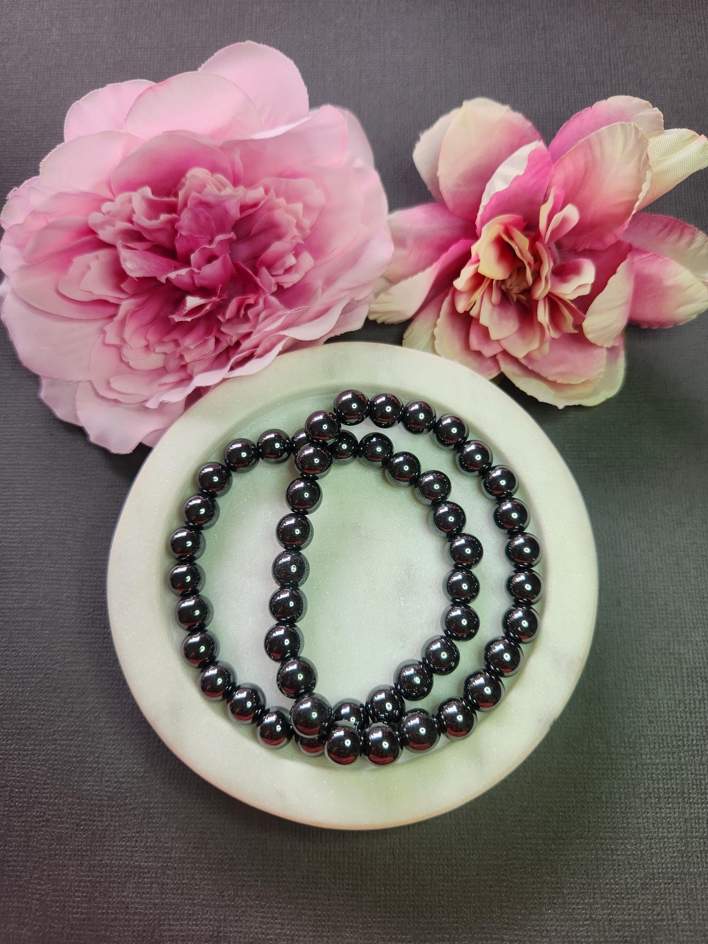 Hematite 8mm beaded bracelets on white dish with two flower props on grey background, natural hematite gemstone beads, handmade crystal jewellery, grounding and protection stone bracelet product photo