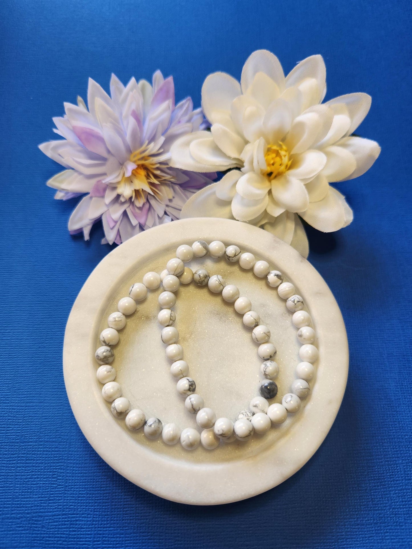 Photo showcasing two Howlite 6mm beaded bracelets displayed in a white dish with two flower props on a blue background, highlighting the polished white crystal beads, calming energy, and stress-relief properties of Howlite — perfect for meditation, mindfulness, and crystal jewelry stacking.