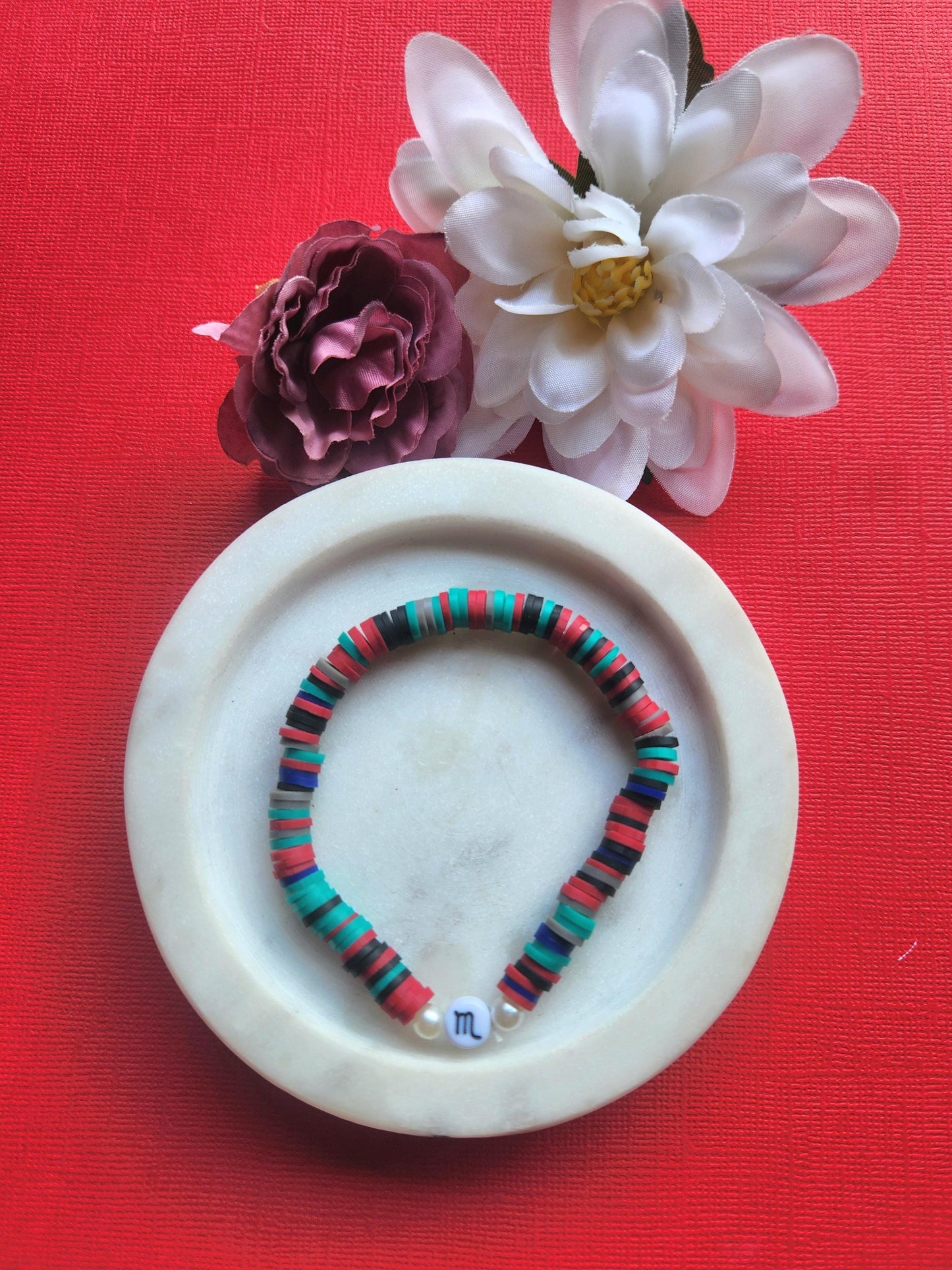 Handmade Scorpio zodiac bracelet displayed in white dish with two flower props against red backdrop, deep teal navy red black clay bead bracelet with Scorpio symbol charm, astrology gift jewellery listing image.