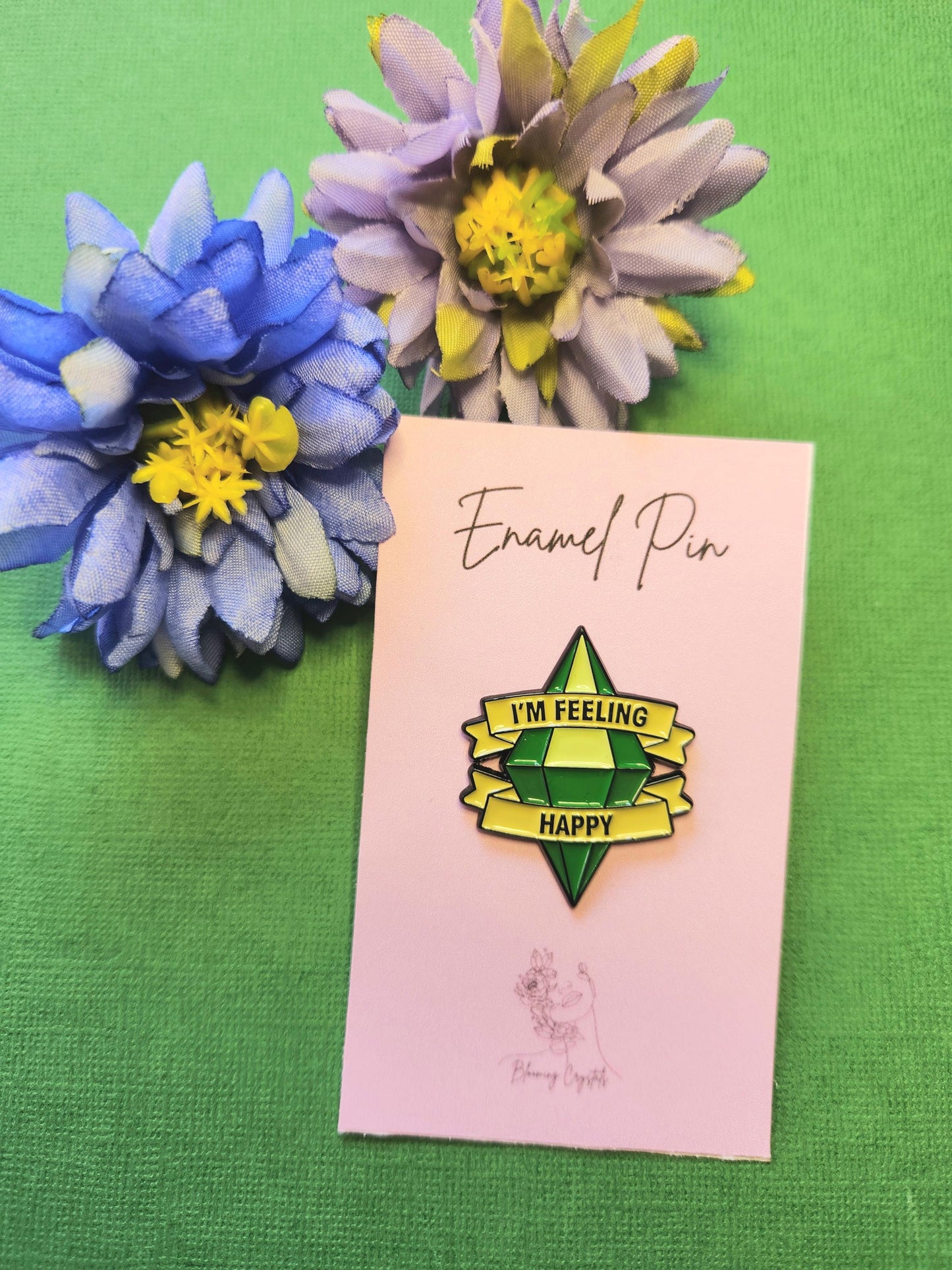 Green diamond-shaped enamel pin with the words “I’m Feeling Lucky,” displayed on a pink backing card with two flower props on a green background, positive affirmation enamel pin, crystal aesthetic pin, cute accessory for bags jackets and gifts.