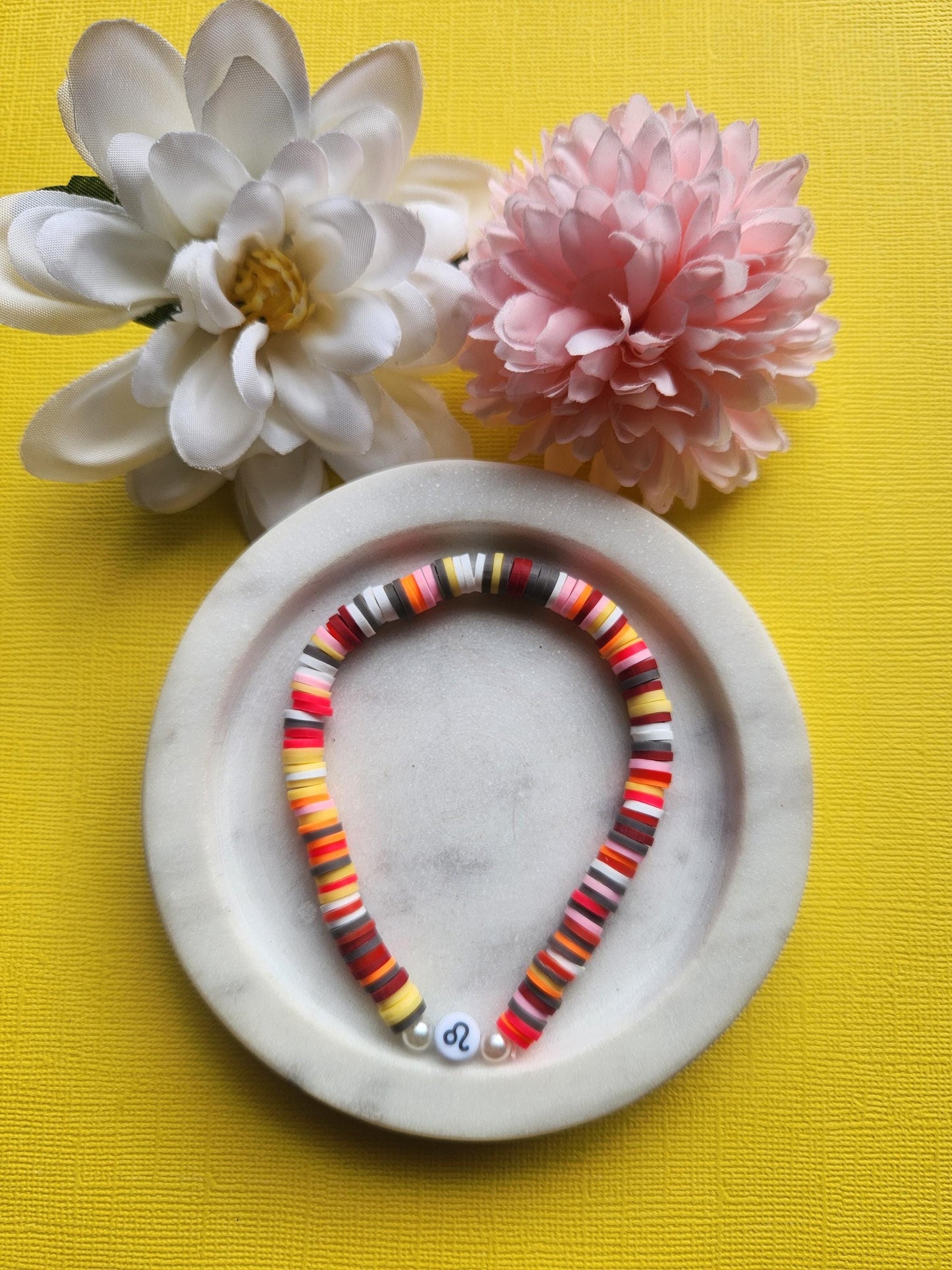 Leo zodiac clay beaded bracelet in white dish with two flower props on yellow background, handmade grey yellow red pink white beaded astrology bracelet with Leo symbol bead, stretchy elastic zodiac jewellery product photo