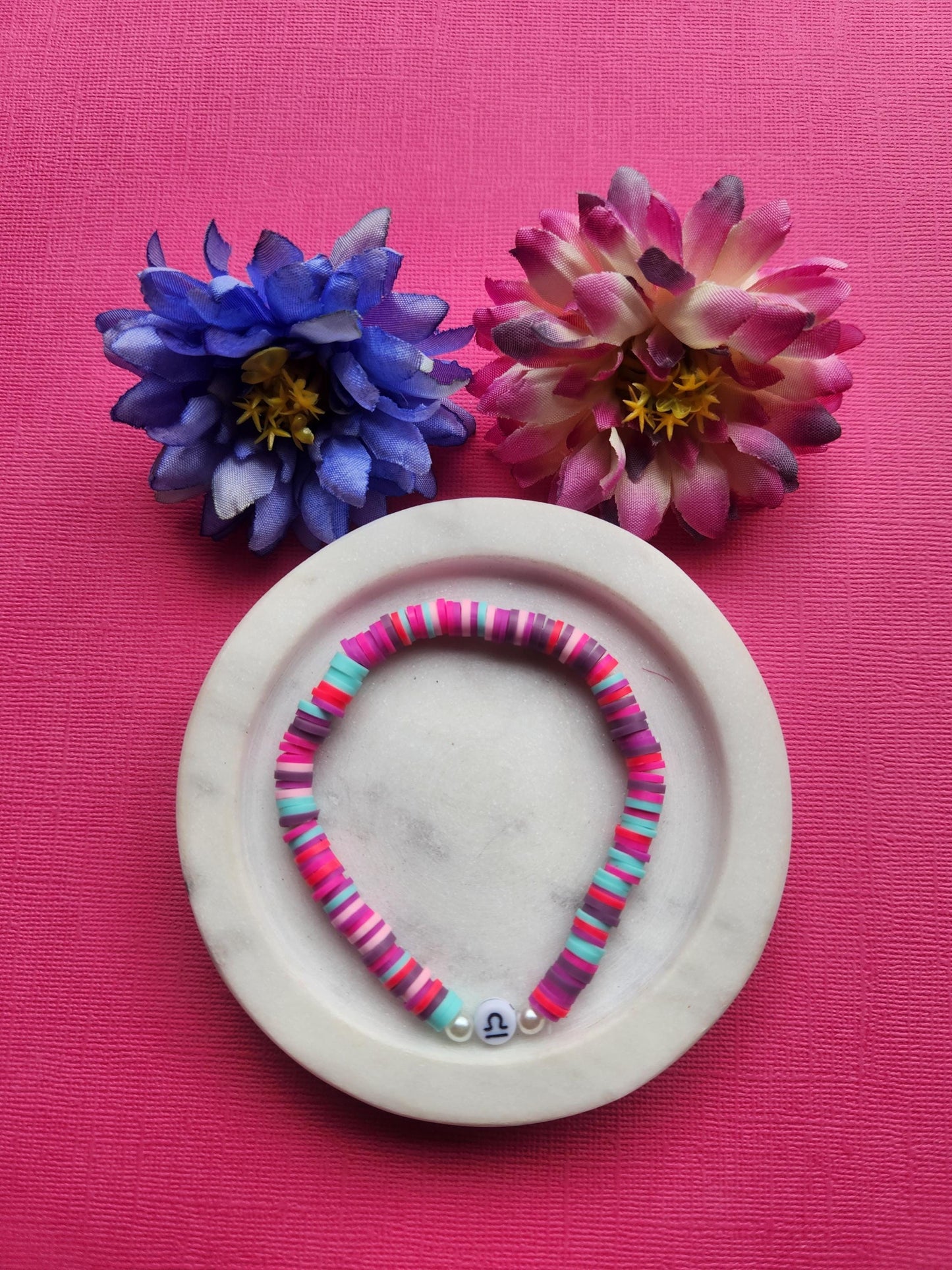 Libra zodiac clay beaded bracelet displayed in a white dish with two flower props on a pink background, colourful handmade astrology bracelet with pink purple light blue red clay beads and Libra symbol bead, stretchy elastic beaded jewellery for Libra gifts, zodiac accessories product photo