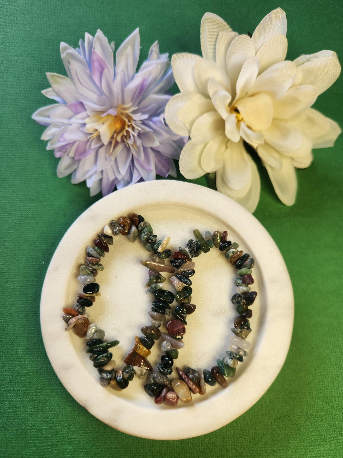 Two Moss Agate crystal chip bracelets displayed in a white dish with two flower props on a bright green background, showcasing natural green tones and polished chip details, perfect for crystal jewelry, grounding energy, and nature-inspired accessories.