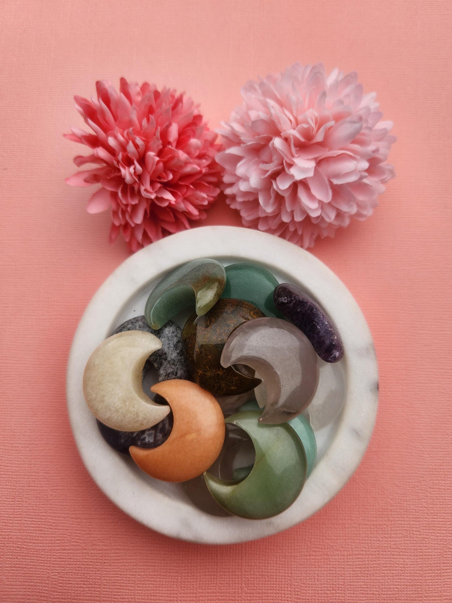 Mystery crystal moon carvings displayed in a white ceramic dish with two floral props on a warm orange background, featuring assorted natural healing crystals and gemstones, crescent moon crystal carvings, metaphysical crystals for intuition manifestation and spiritual energy, small crystal moons perfect for gifting altar decor and crystal collections