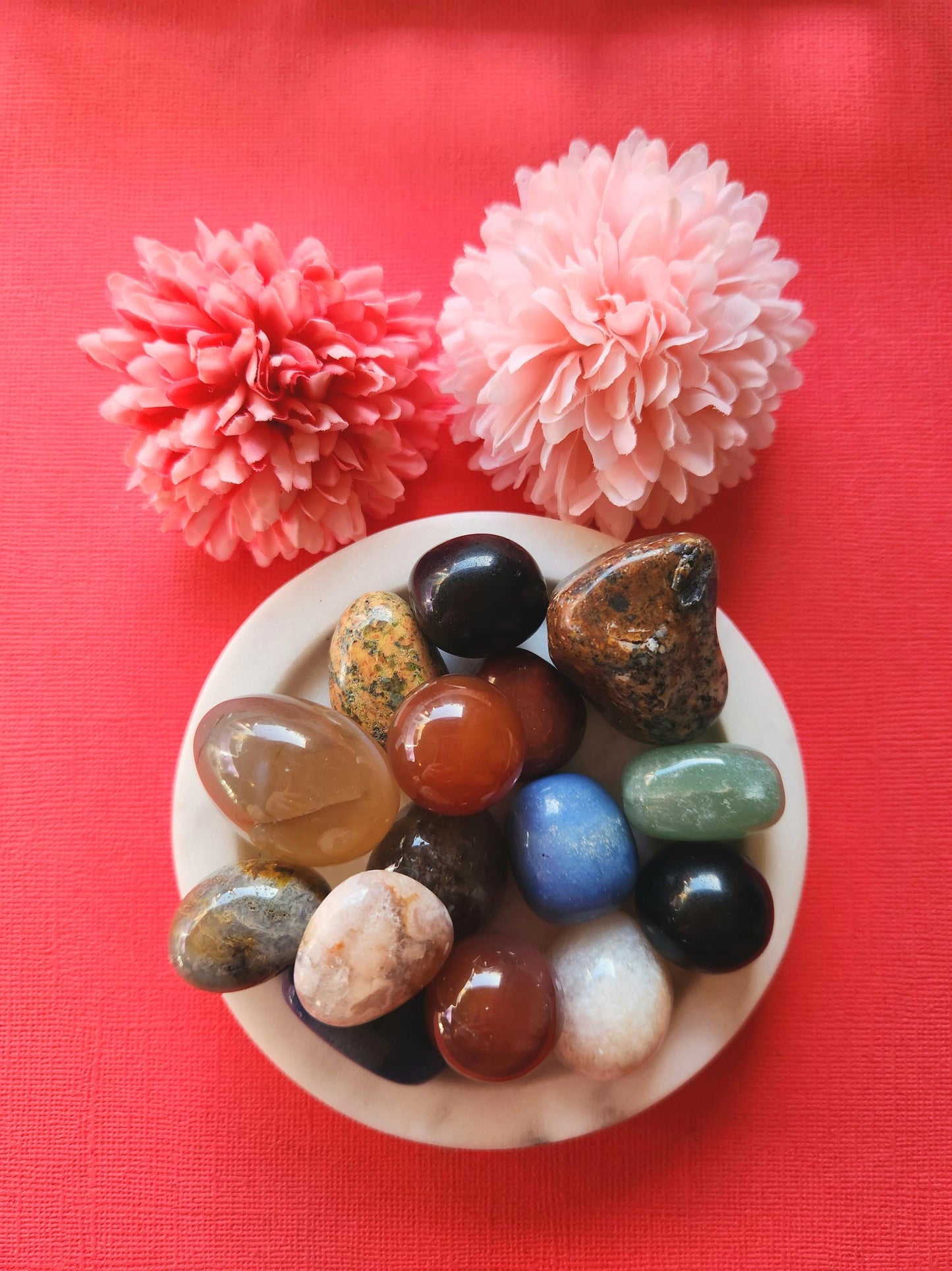 A selection of mystery crystal tumbles displayed on a vibrant red background. Each polished gemstone is intuitively chosen, offering a unique surprise for crystal lovers. These tumbled stones may include a variety of healing crystals for energy work, meditation, or gifting. The bold red backdrop adds excitement and contrast, emphasizing the vibrant and varied colors of the mystery stones.