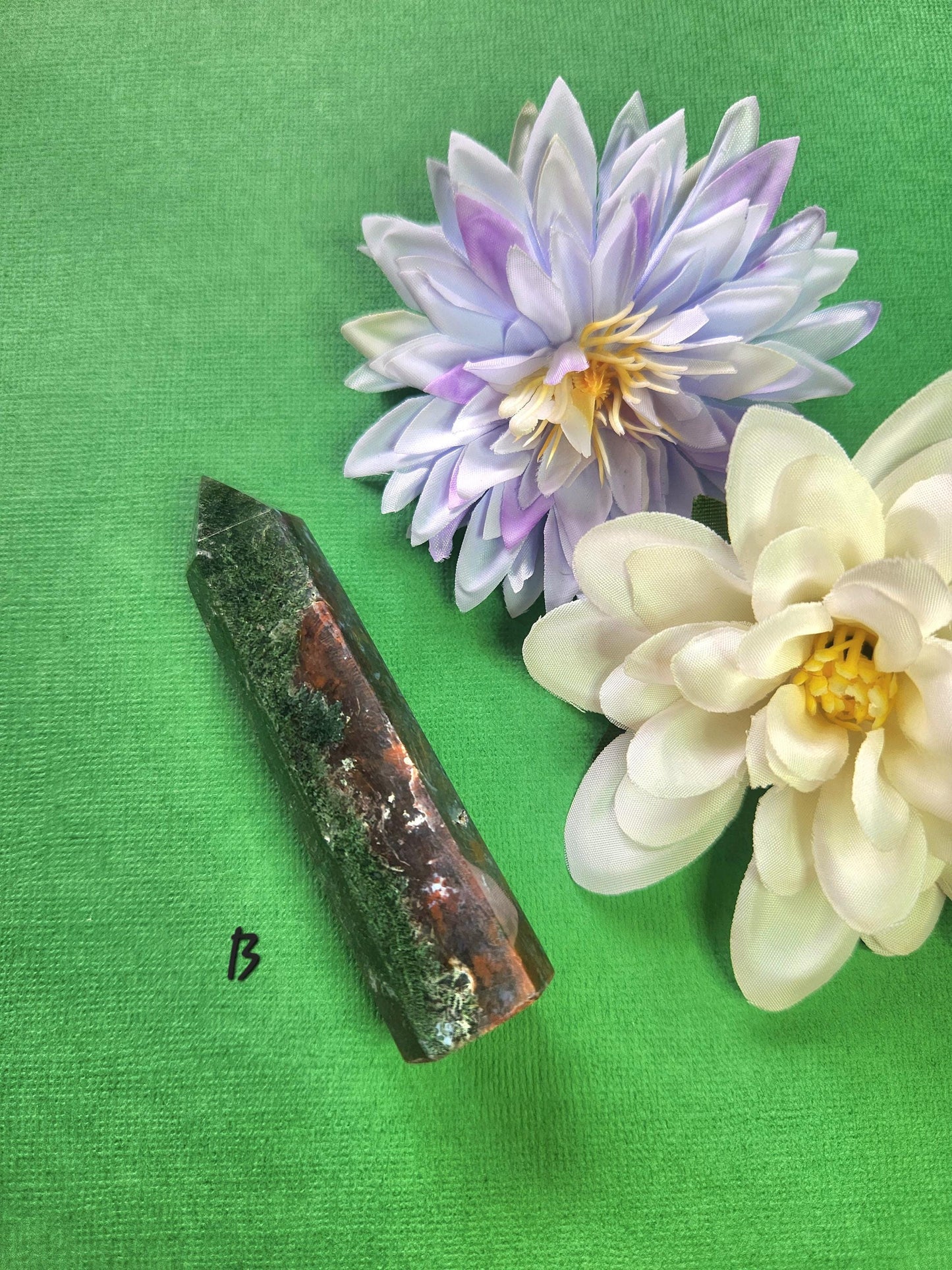 Polished Ocean Jasper tower B showing unique green, cream, and brown orbicular formations, styled with two flower props on a green background, premium healing gemstone for grounding, heart chakra support, metaphysical décor, and spiritual crystal use