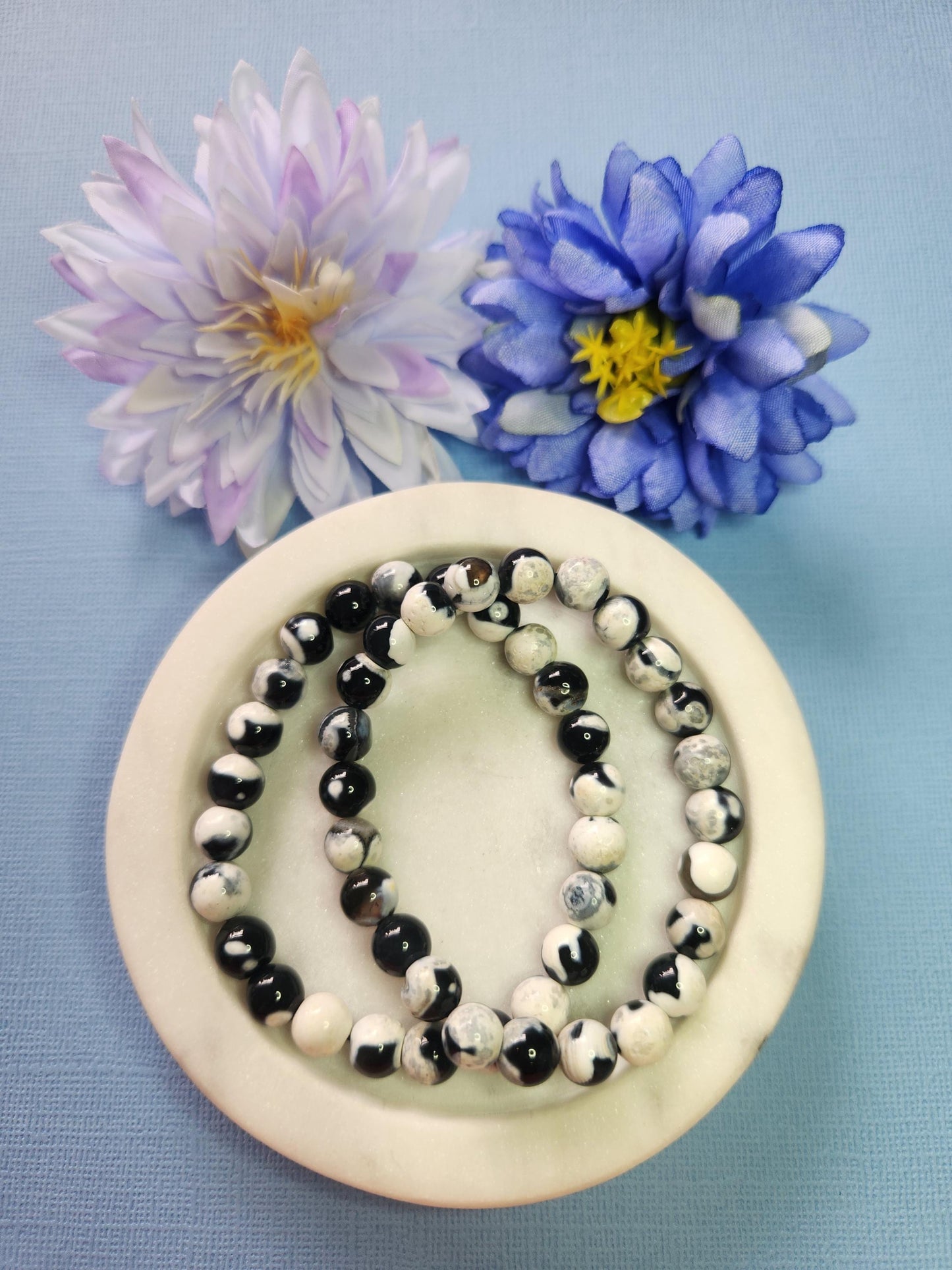 Orca Agate 8mm beaded bracelets on white dish with two flower props on blue background, natural orca agate gemstone beads, handmade crystal jewellery, calming protection stone bracelet product photo