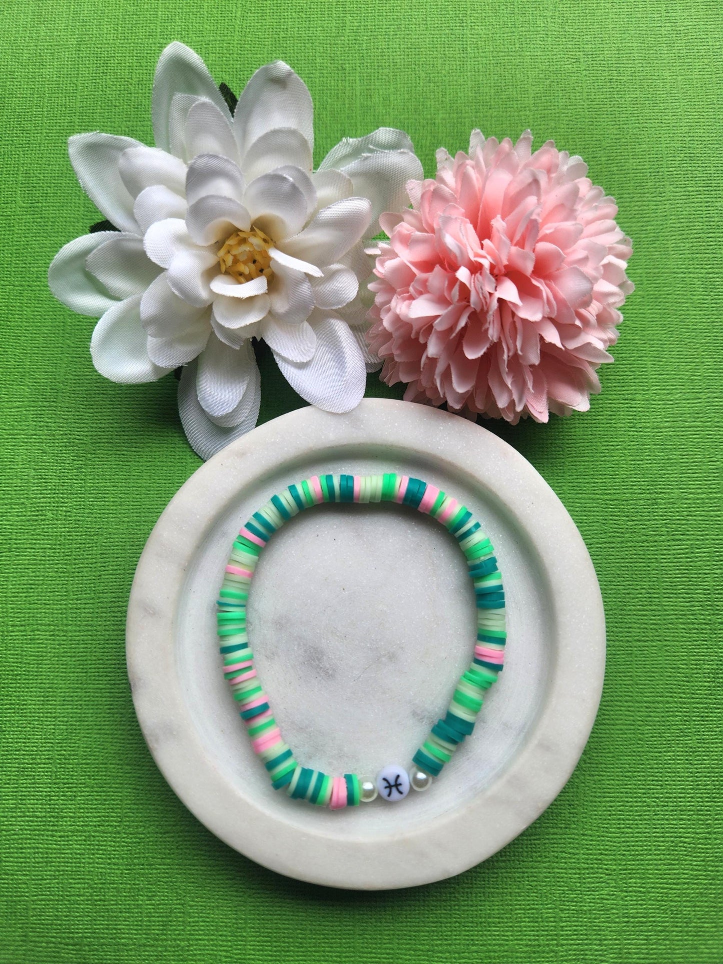 Pisces Zodiac clay beaded bracelet with mint, green, dark green, and light pink beads, featuring two faux pearl accent beads framing the Pisces charm, displayed on a white dish with flowers on green background — handmade astrology jewelry, horoscope gift, crystal-inspired accessory.