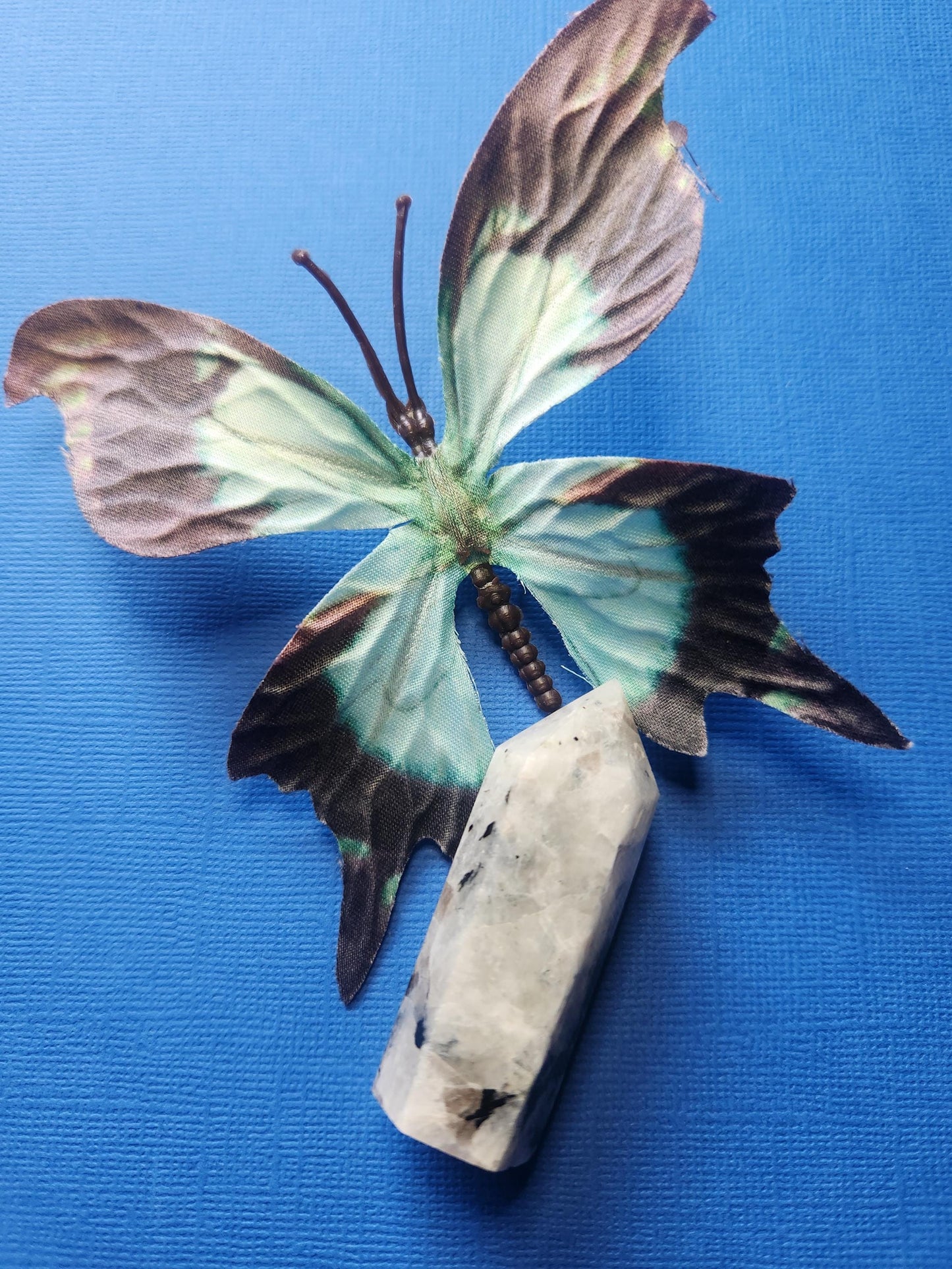 Rainbow moonstone crystal tower showing its opposite side, displayed with a blue butterfly prop against a dark blue background. Hand-carved and polished, this tower enhances intuition, emotional balance, and spiritual growth — ideal for meditation, crystal collections, and home décor.