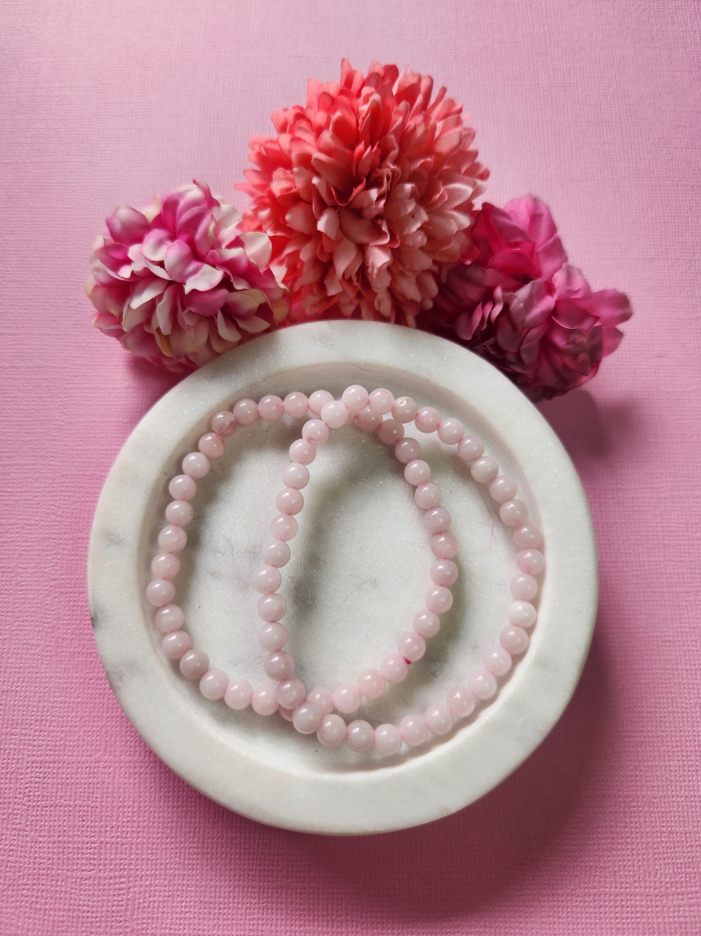 Rose Quartz 6mm beaded crystal bracelets displayed in a white dish with two flower props on a light pink background, featuring natural soft pink gemstone beads for love, compassion, and heart chakra healing crystal jewelry photography.