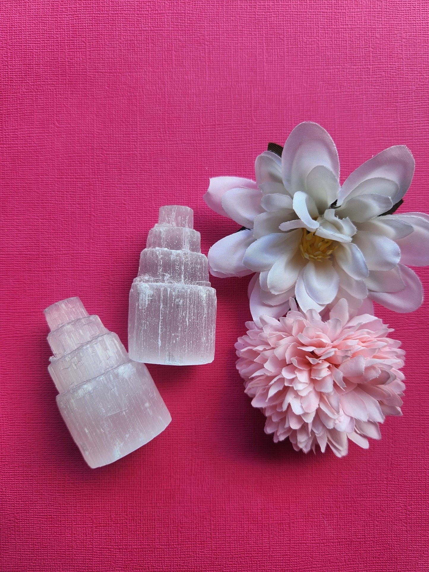 Pair of selenite crystal towers displayed upright on a pink background with delicate flower props featuring polished white selenite points for crystal decor healing meditation and metaphysical energy