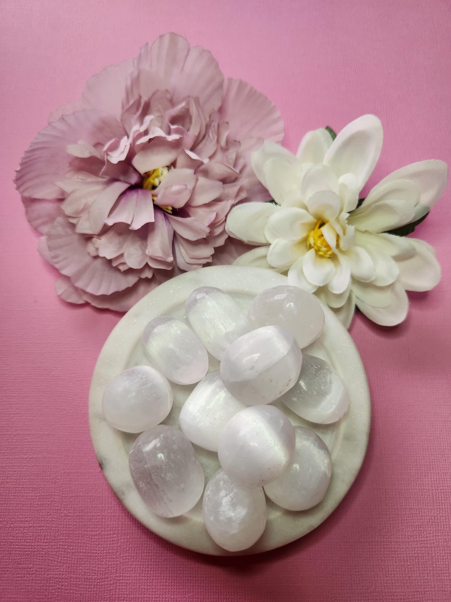 Close-up photo of several selenite tumbles displayed in a white dish with two soft flower props on a pink backdrop. The tumbles show translucent white hues with a smooth, polished finish. Ideal for selenite crystals, tumbled gemstones, crystal collections, and decorative crystal accents