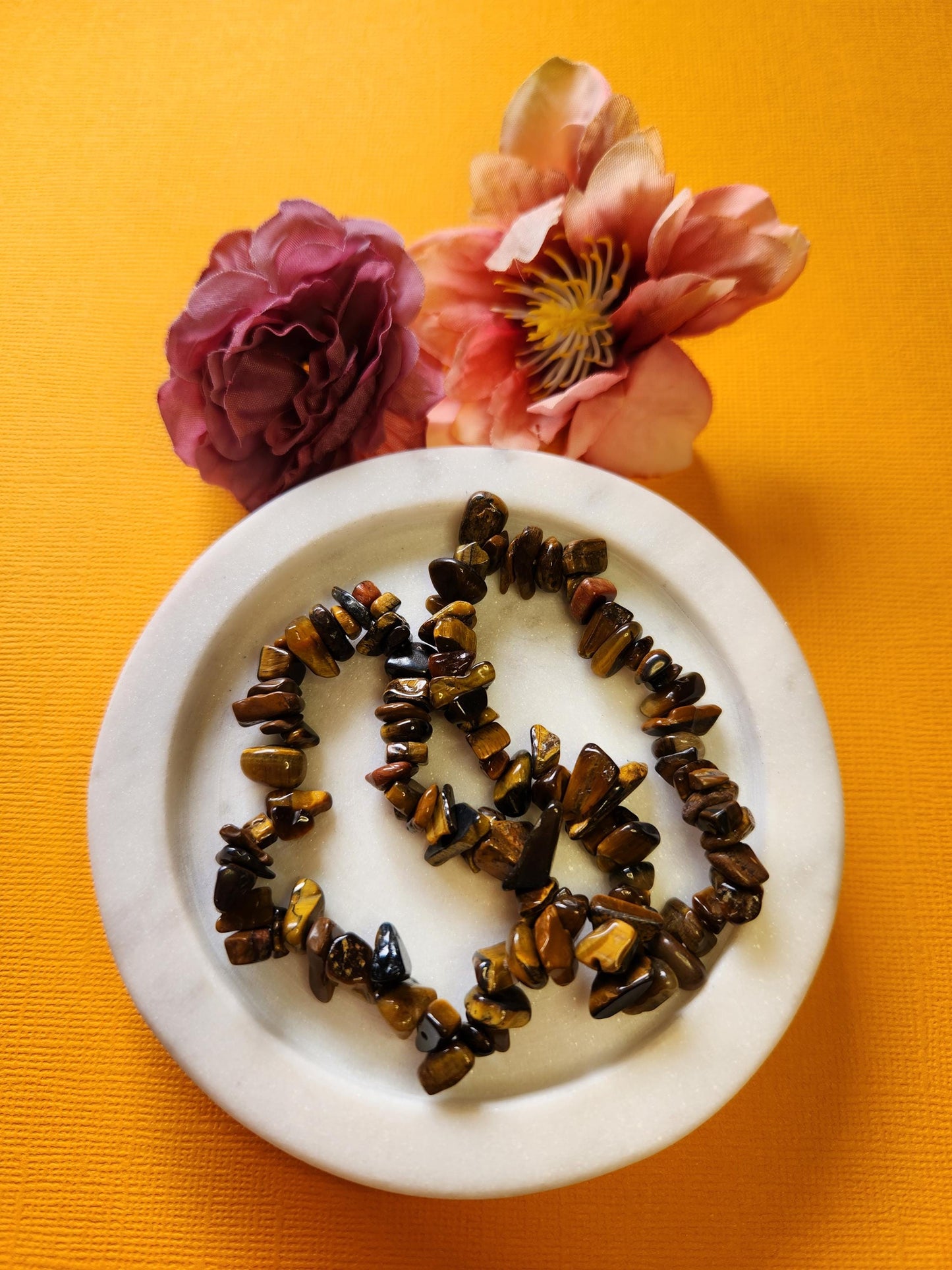 Two Tigers Eye chip bracelets on a white dish with two flower props, yellow background, natural gemstone, healing crystal, meditation jewelry.
