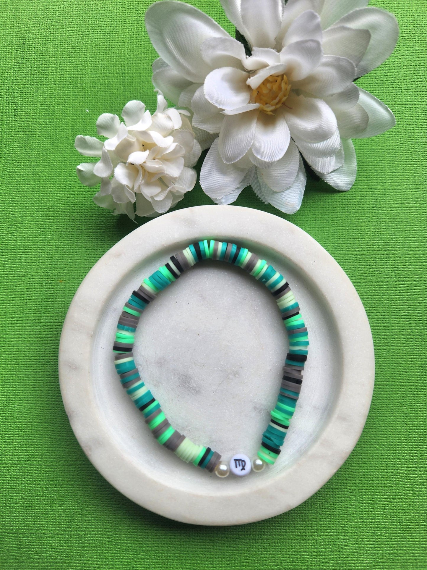 Virgo zodiac clay beaded bracelet displayed in a white dish on a green background with two flower props, handmade elastic bracelet featuring green, black and brown polymer clay beads with a plastic Virgo symbol bead, astrology jewellery for Virgo gifts, zodiac accessories, handmade beaded bracelets, spiritual gift ideas and earthy aesthetic jewellery