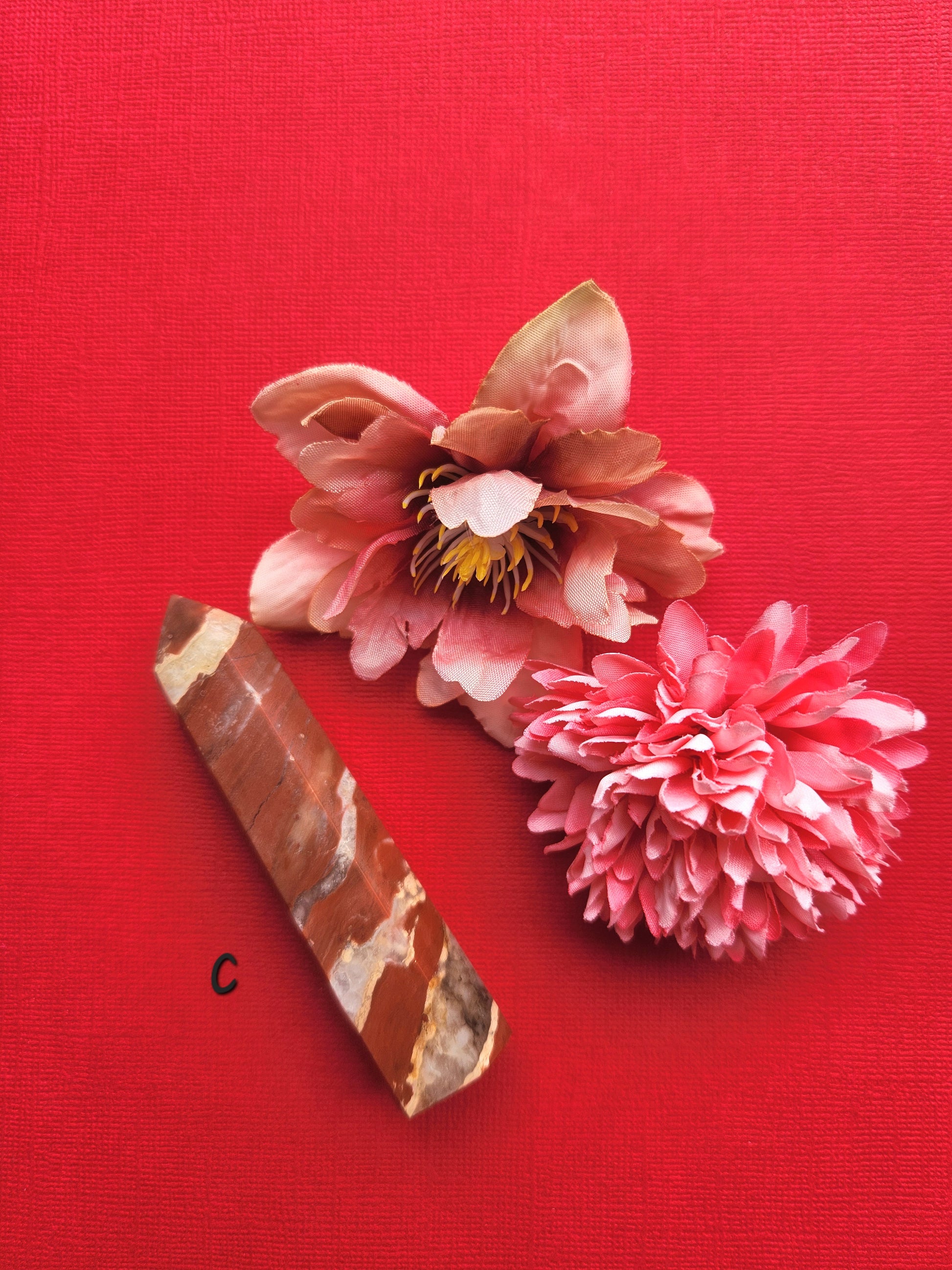 Polished Red Jasper crystal tower (Tower C) displayed on a red background with two decorative flower props, showing its deep red color, natural patterns, and grounding energy. Perfect for meditation, Reiki, crystal healing, spiritual decor, and home altars.