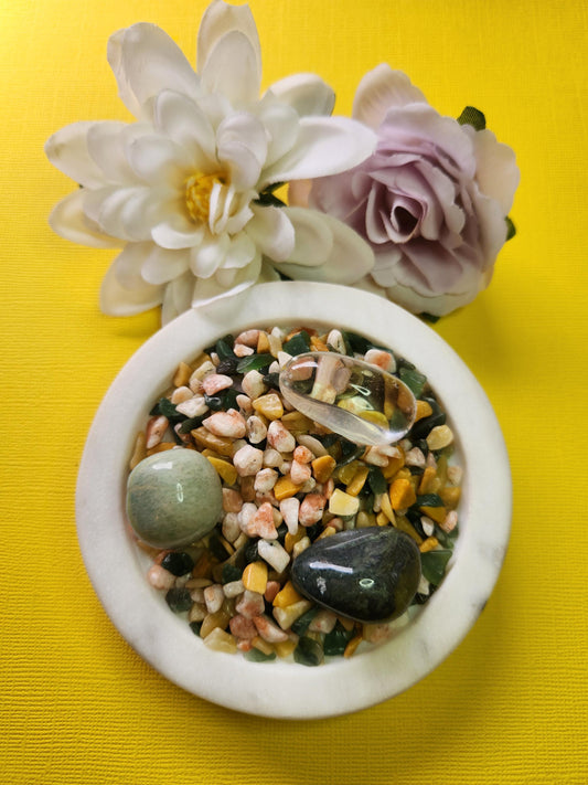 Abundance Spell Confetti featuring crystal chips of Yellow Aventurine, Sunstone, and Green Aventurine, with tumbled stones of Moss Agate, Clear Quartz, and Amazonite. The 50g crystal mix sits beautifully on a white dish with two flower props against a vibrant yellow background, radiating uplifting, wealth-attracting, and prosperity energy. Perfect for abundance rituals, crystal magic, manifestation grids, and spiritual decor.