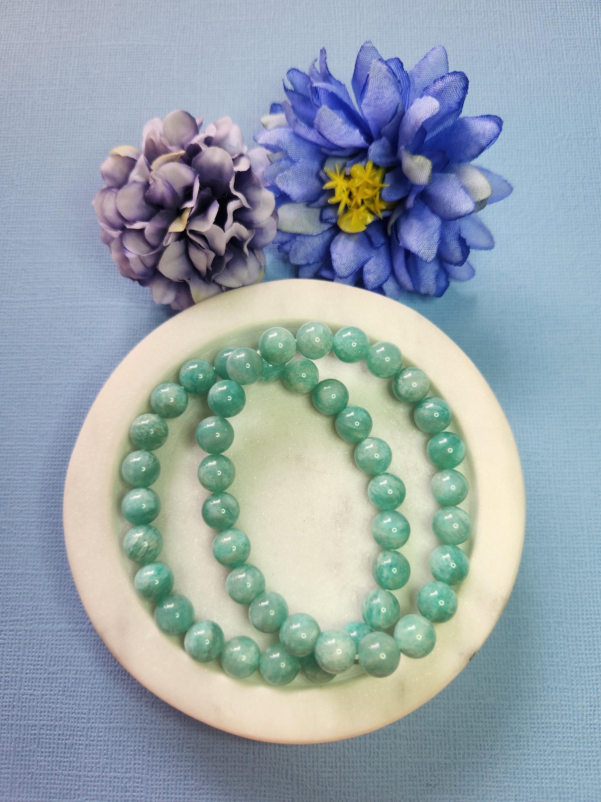 Amazonite 8mm beaded bracelet in white dish with two flower props on blue background, natural amazonite gemstone beads, handmade crystal jewellery, calming soothing communication stone bracelet product photo
