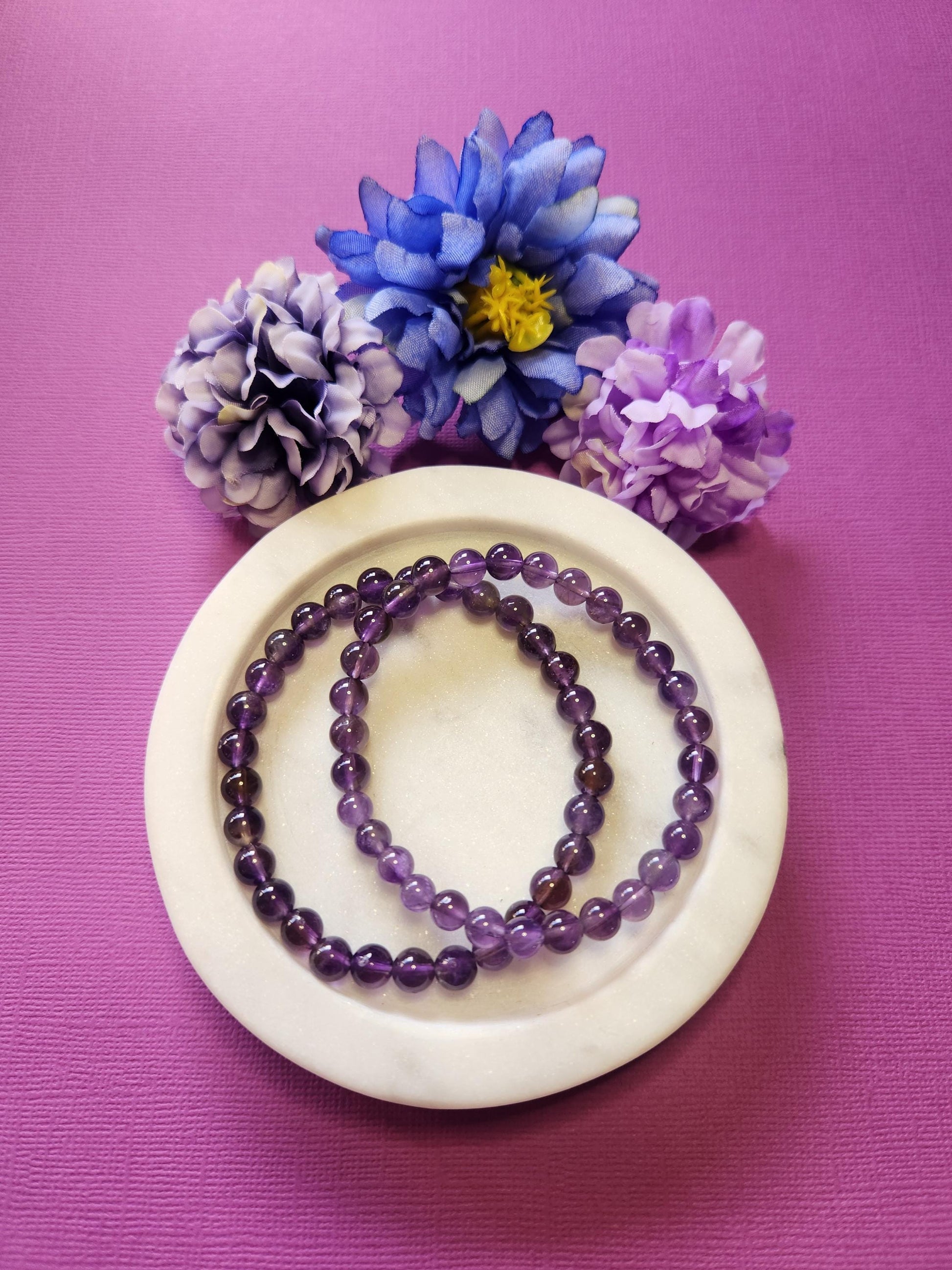 Photo showcasing two 6mm Amethyst crystal beaded bracelets in a white dish with three flower props on a purple background, highlighting the polished purple quartz beads, spiritual healing energy, and calming properties of Amethyst — perfect crystal jewelry for meditation, intuition, and stress relief.