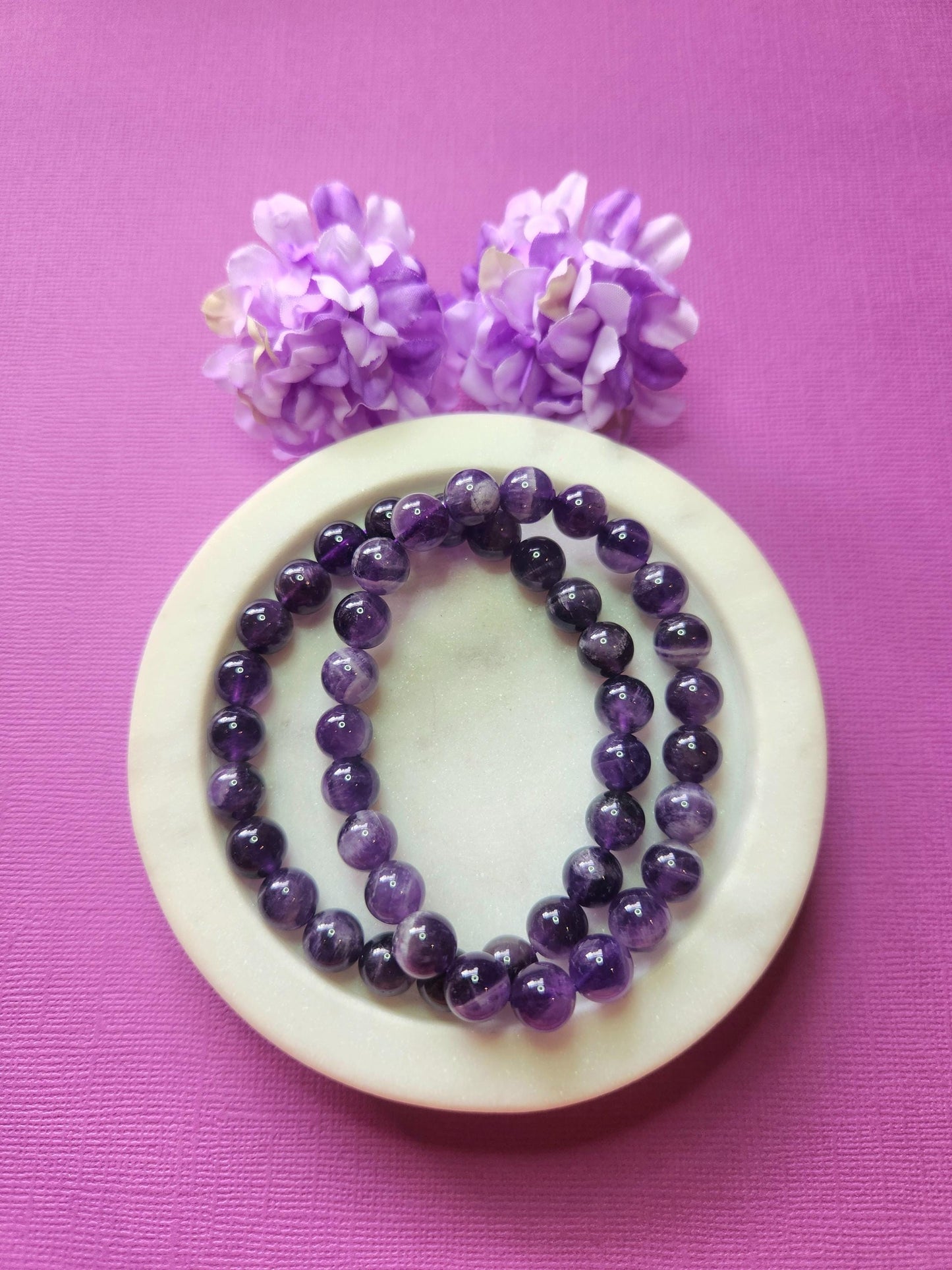 Two Amethyst 8mm beaded bracelets in white dish with two flower props on purple background, natural amethyst gemstone beads, handmade crystal jewellery, calming, spiritual and protective stone bracelet product photo