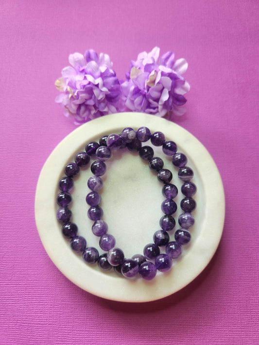 Two Amethyst 8mm beaded bracelets in white dish with two flower props on purple background, natural amethyst gemstone beads, handmade crystal jewellery, calming, spiritual and protective stone bracelet product photo
