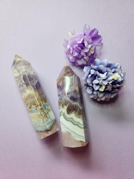 Two Amethyst and Crazy Lace Agate Crystal Towers displayed next to two flower props on a lavender background. Polished gemstone towers with natural purple amethyst and vibrant orange crazy lace agate patterns. Healing crystal towers for meditation, chakra balancing, energy cleansing, spiritual décor, and crystal collections.