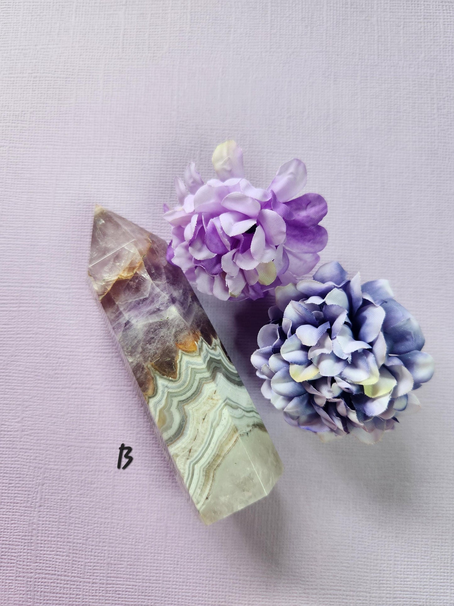 Close-up of Tower B: Amethyst and Crazy Lace Agate Crystal Tower on a lavender background with two flower props. Polished natural gemstone tower with rich purple amethyst and vibrant orange crazy lace agate patterns. Perfect for meditation, chakra healing, energy balancing, spiritual décor, and crystal collections.