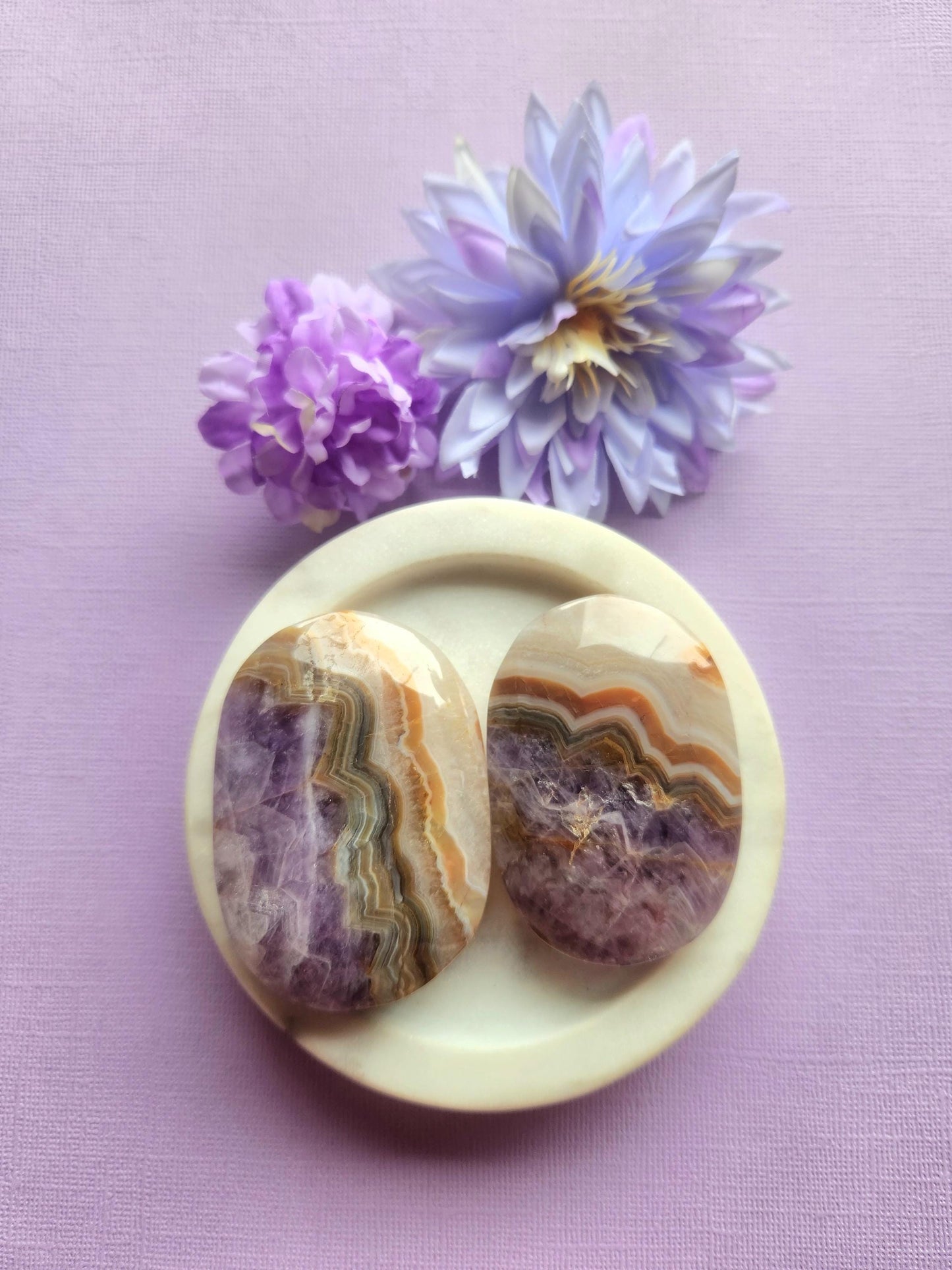 Amethyst and crazy lace agate palmstones displayed in a white ceramic dish with two floral props on a soft lavender background, featuring polished natural crystal palm stones, healing gemstones for calm balance grounding and joy, metaphysical crystals perfect for meditation gifting altar decor and crystal collections