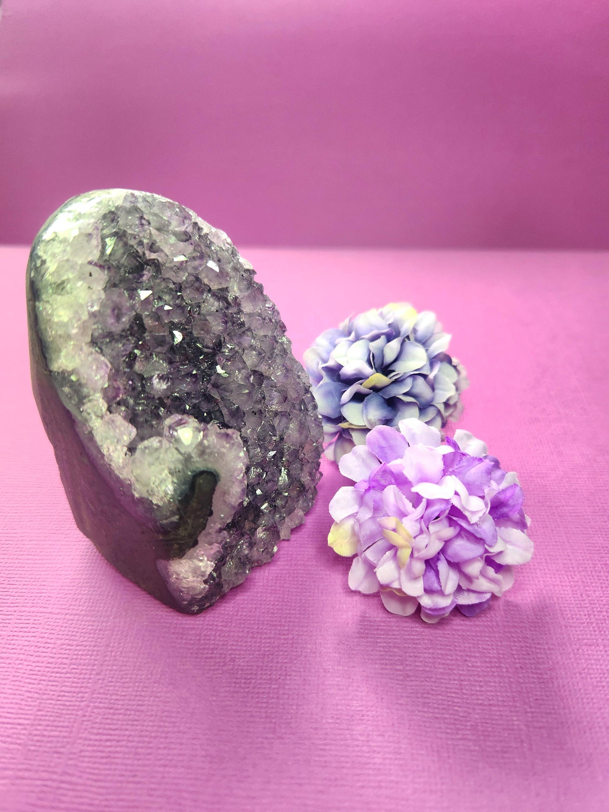 Side view of Amethyst Cluster Cut Base on a purple background with two flower props. Raw amethyst geode showing natural crystal points and polished flat base. Purple healing crystal for calm, protection, and spiritual energy cleansing. Perfect décor piece for meditation or crystal collections.