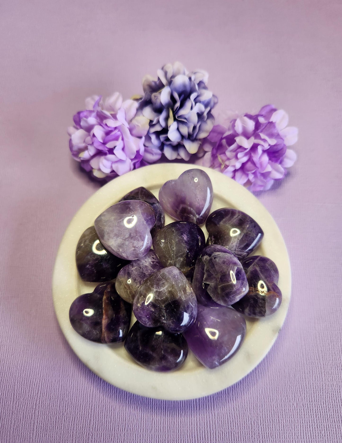 Amethyst crystal heart stones displayed in a white dish on a purple background with flower props featuring polished purple amethyst hearts for calming stress relief spiritual healing meditation crystal decor and gift