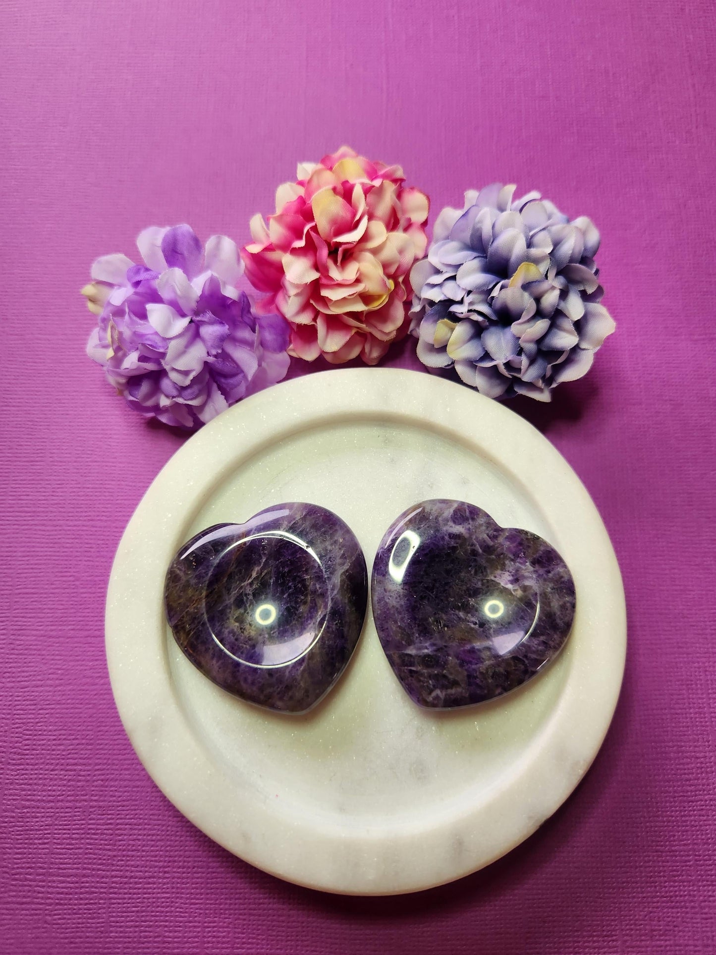 Two Amethyst heart-shaped worry stones displayed in a white dish with two delicate flower props on a soft purple background; smooth polished purple crystal hearts for calming and stress relief.